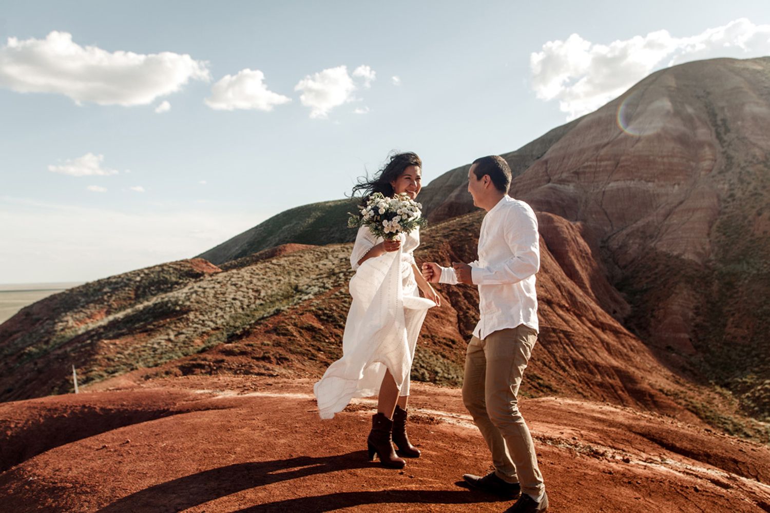 Couple in white outfits walking on red volcanic terrain with mountains and blue sky in the background.