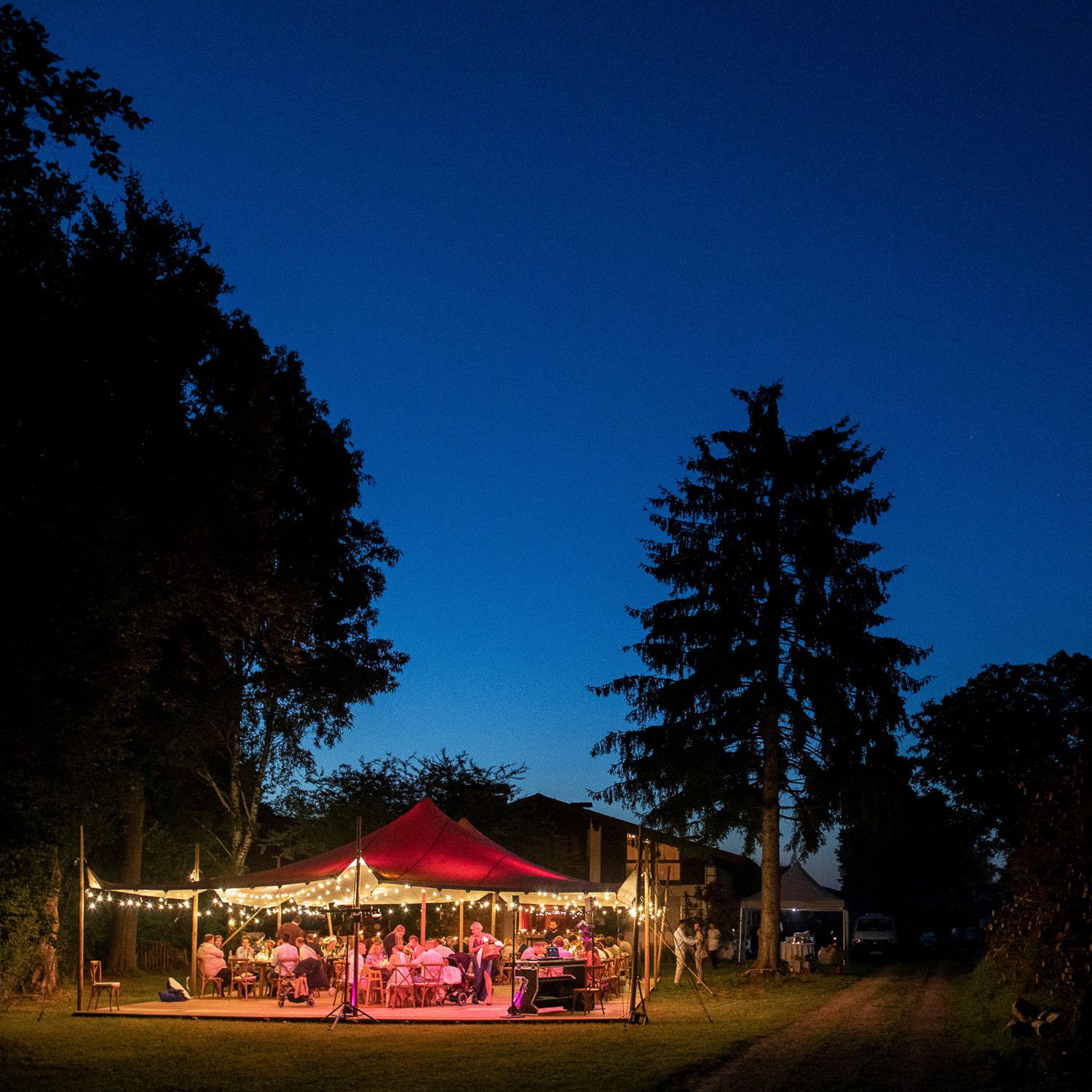 Illuminated red tent canopy glows at dusk with string lights creating a magical outdoor event setting in a wooded area.