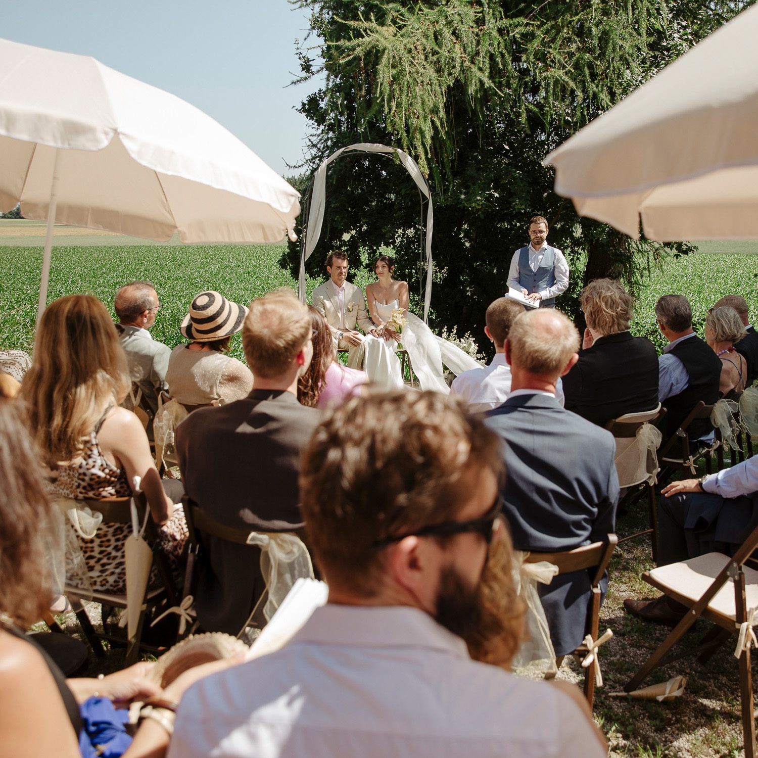 Outdoor wedding ceremony with guests seated under white umbrellas on a sunny summer day.