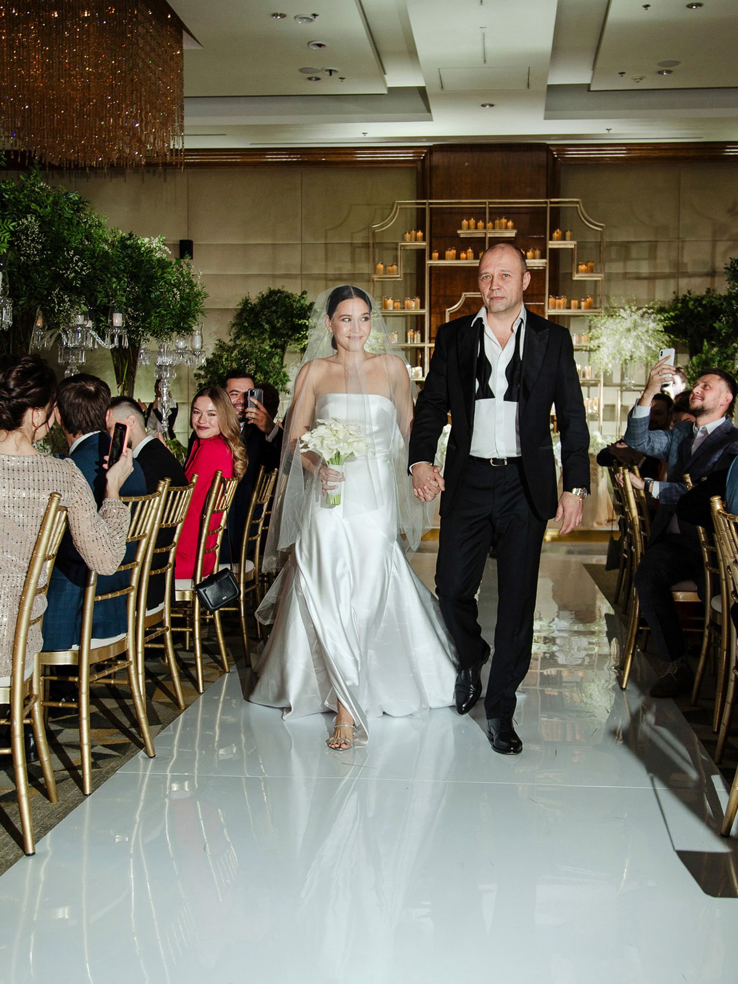 Wedding guests enjoying a formal reception in an elegant ballroom with gold chairs and crystal chandeliers.
