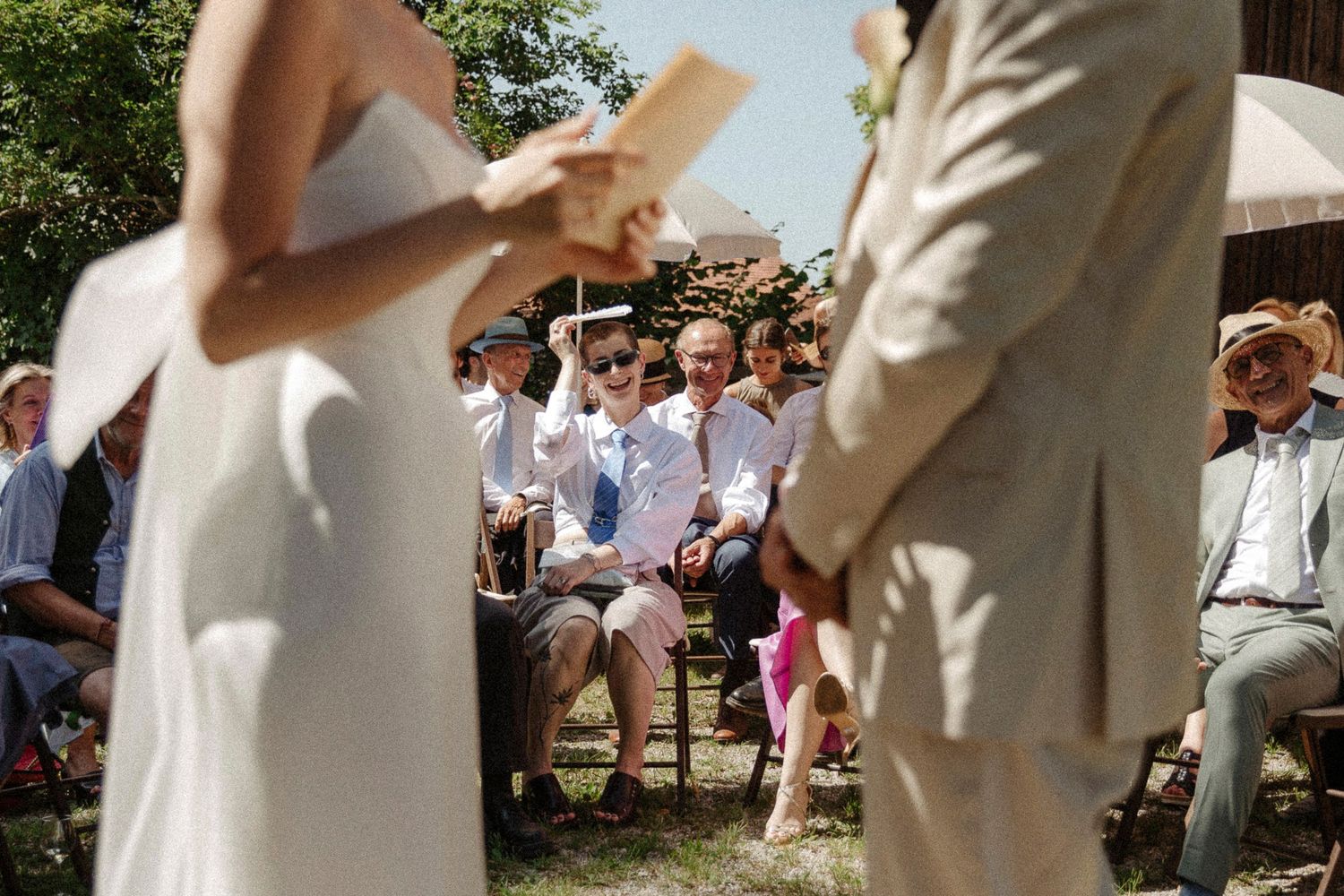 A group of wedding guests gather outdoors on a sunny day to celebrate and throw confetti in the air.