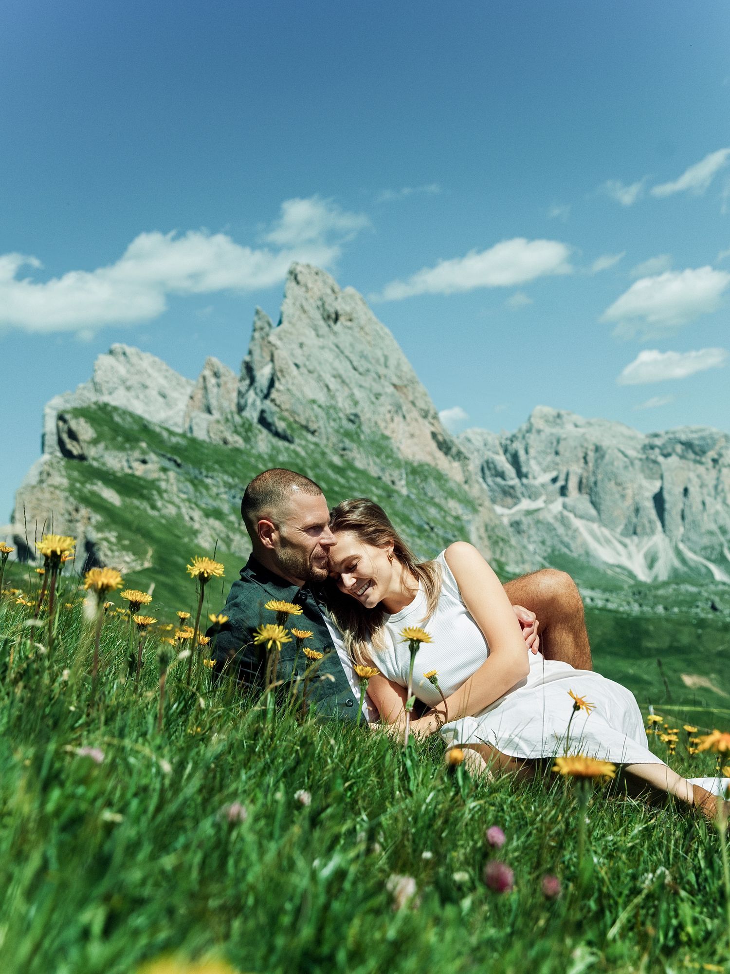 Couple sitting in a green meadow with yellow wildflowers, embracing with rocky mountain peaks under blue sky.