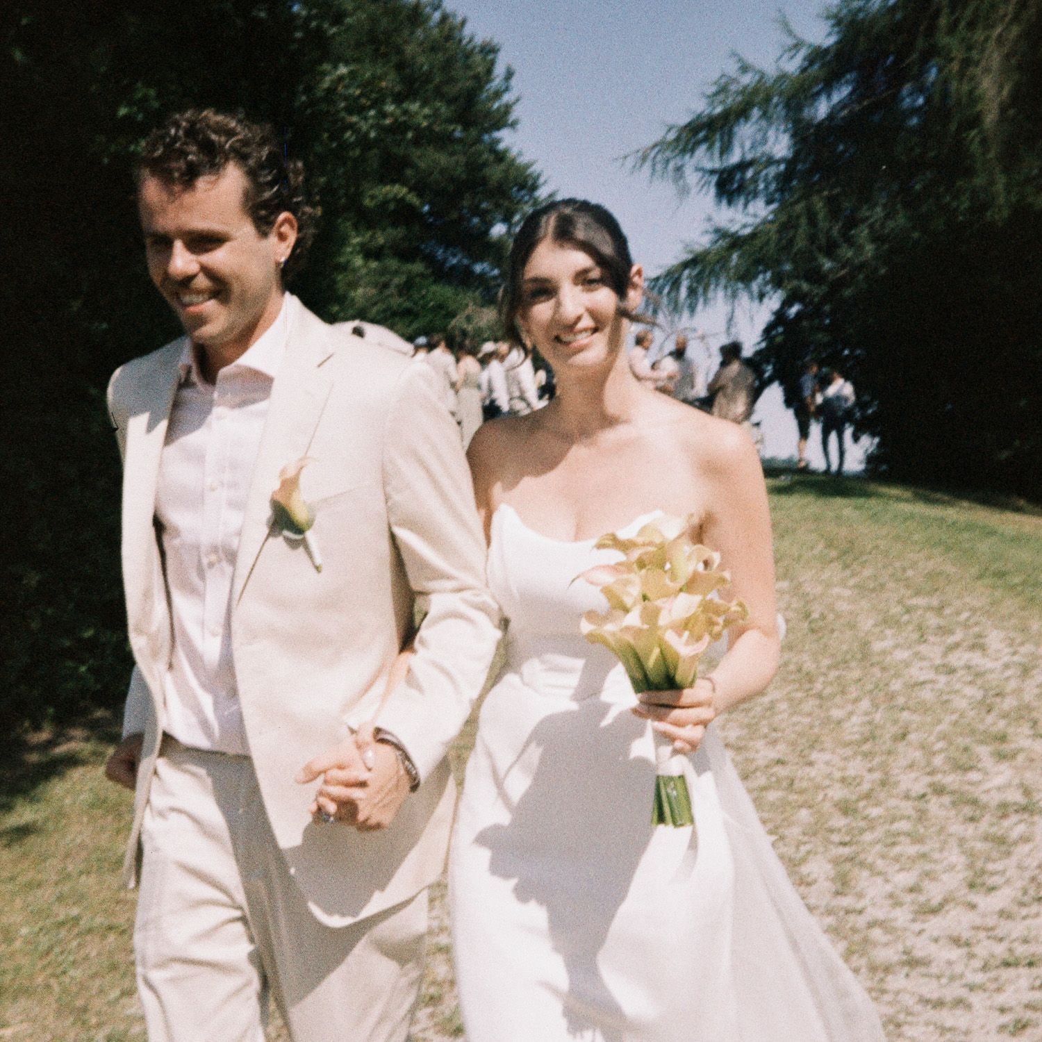 A couple in wedding attire walks together on a sunny beach, their white outfits glowing against the sand and sea.
