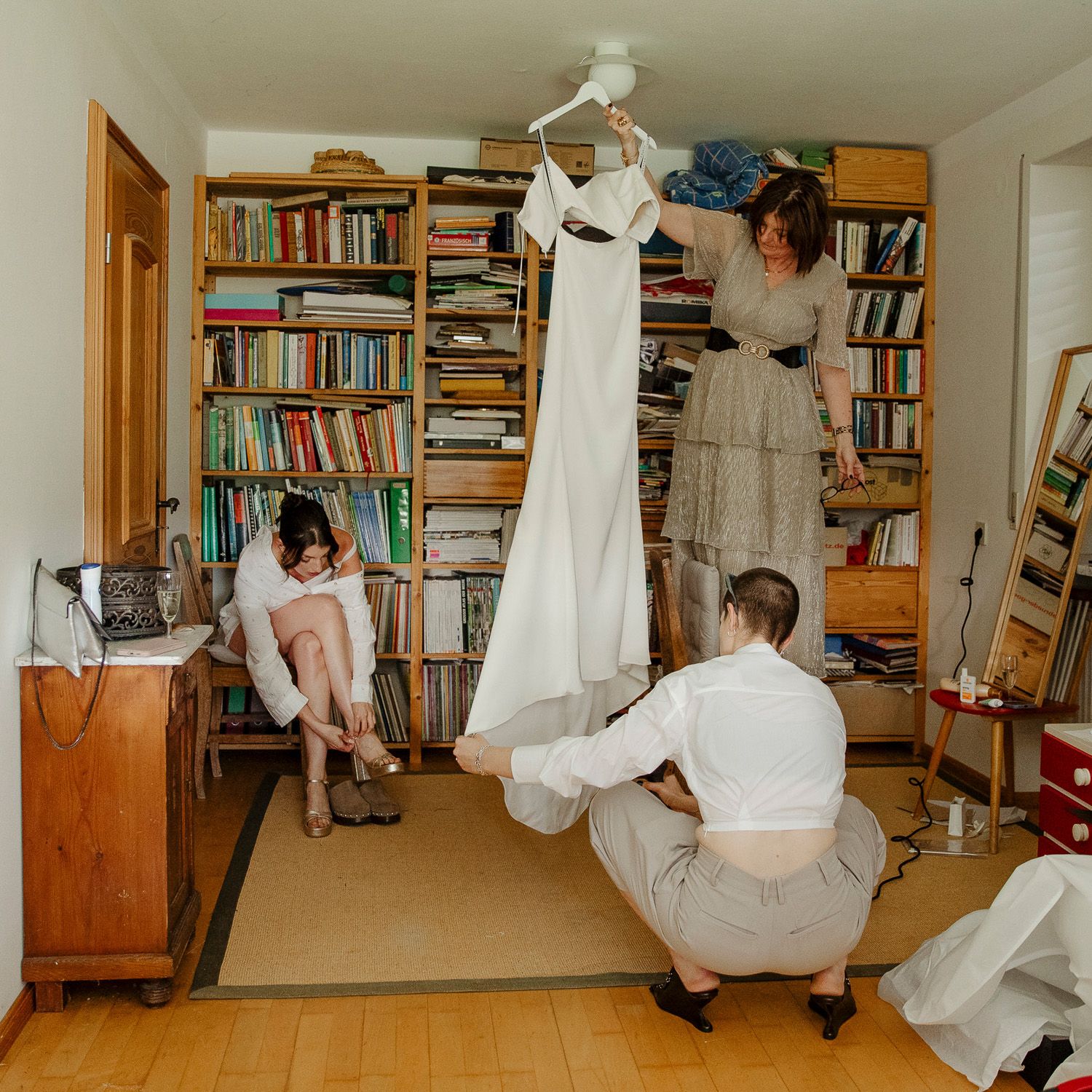 A wedding dress hangs in a cozy room with floor-to-ceiling bookshelves while people prepare for a special day.