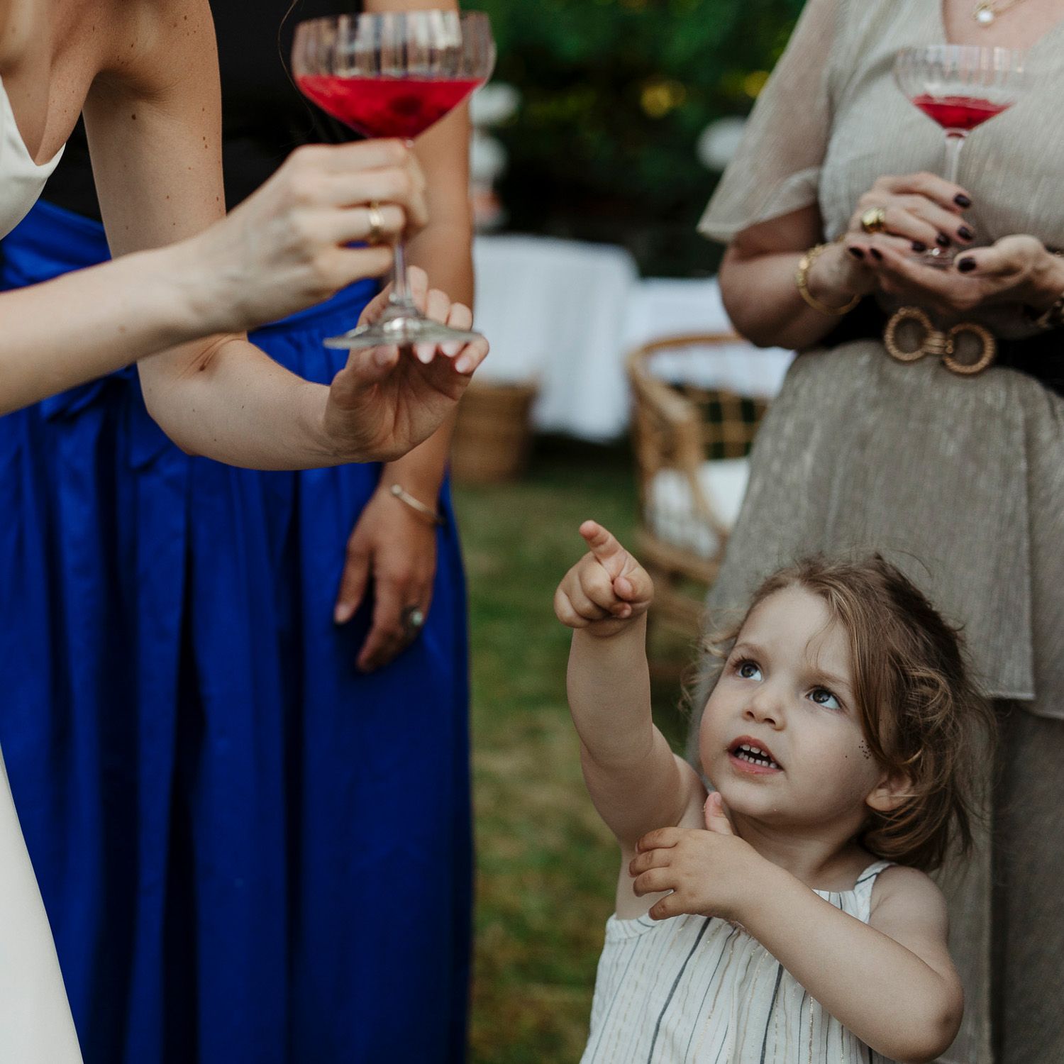 A child in a white dress reaches up towards bubbles floating in the air at an outdoor event with people in blue dresses.