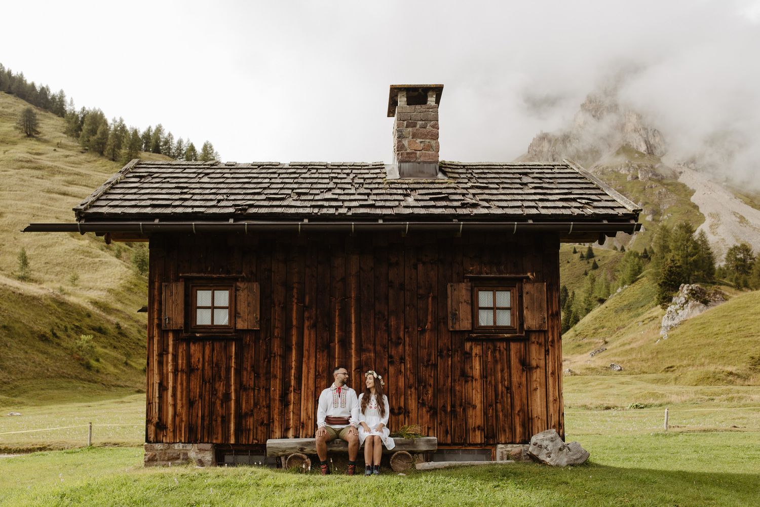 Two people sit together on the porch of a rustic wooden cabin in the misty Alps mountains.