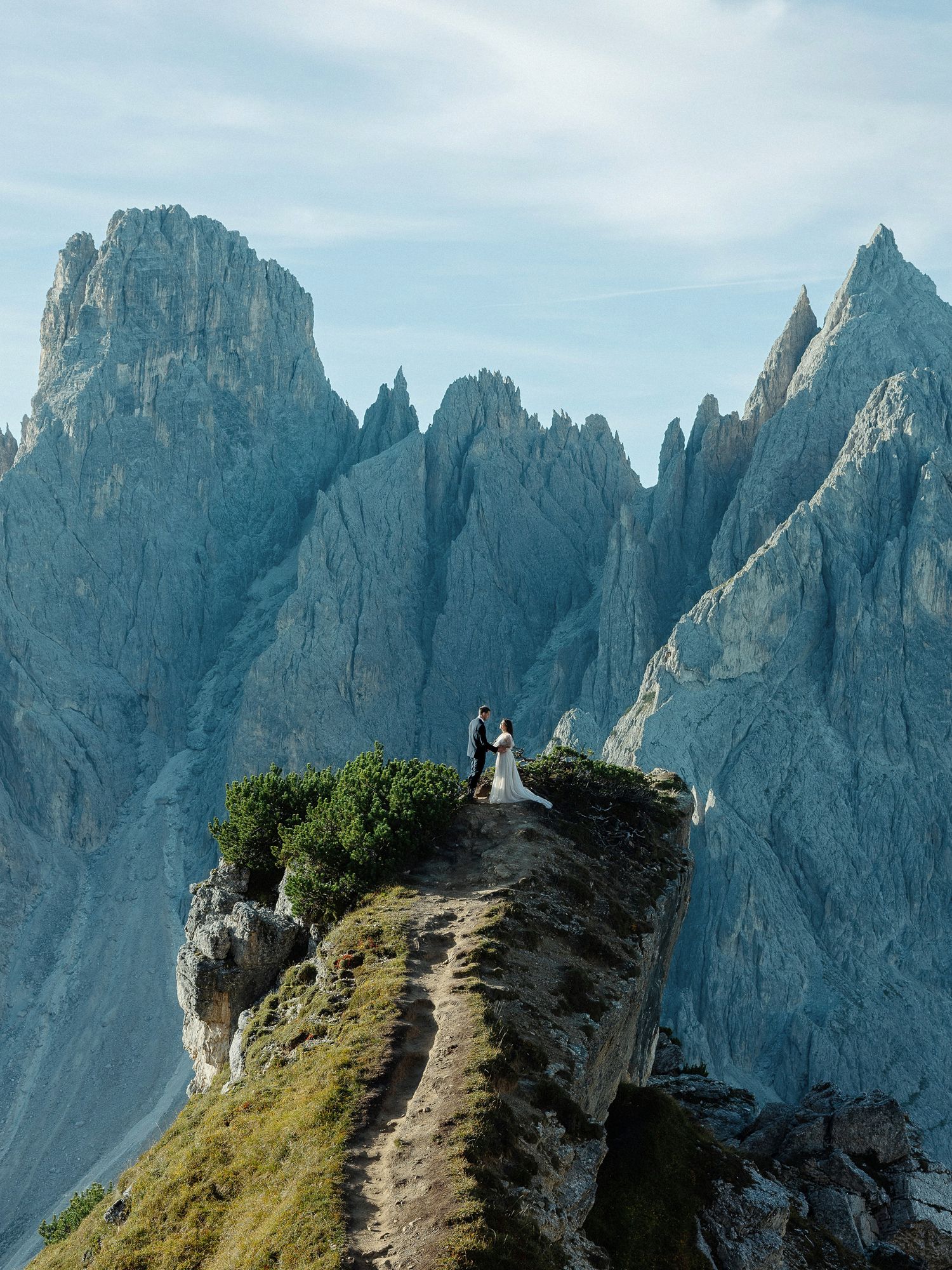 Couple stands on a rocky mountain peak in the Dolomites, surrounded by dramatic jagged peaks under a blue sky.