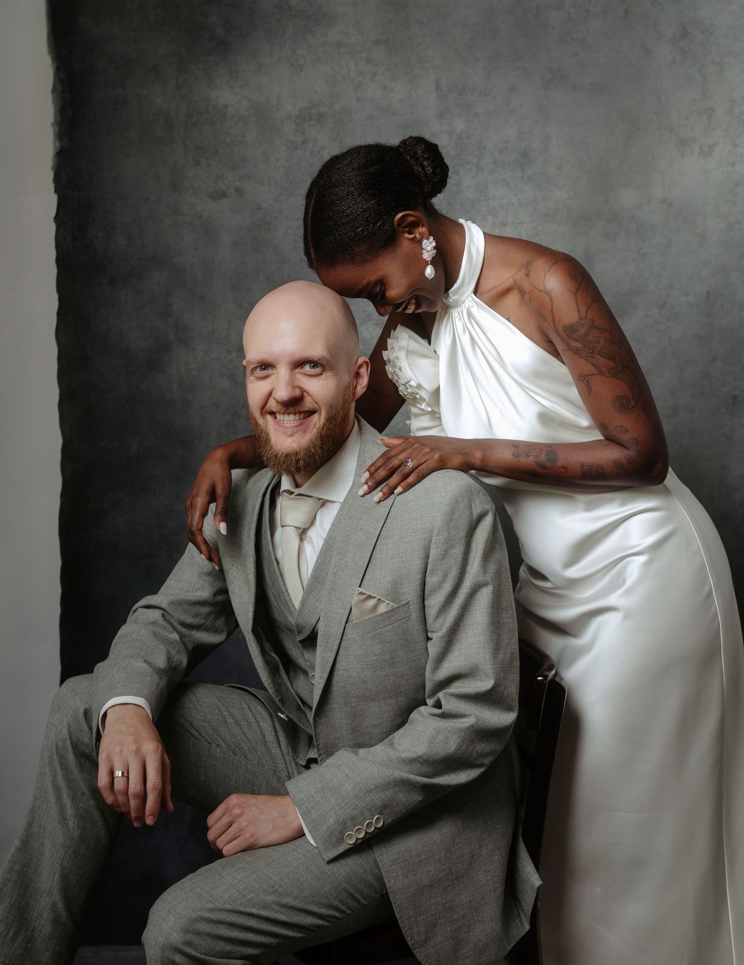 A couple shares an intimate moment in formal attire during a portrait session against a gray backdrop.