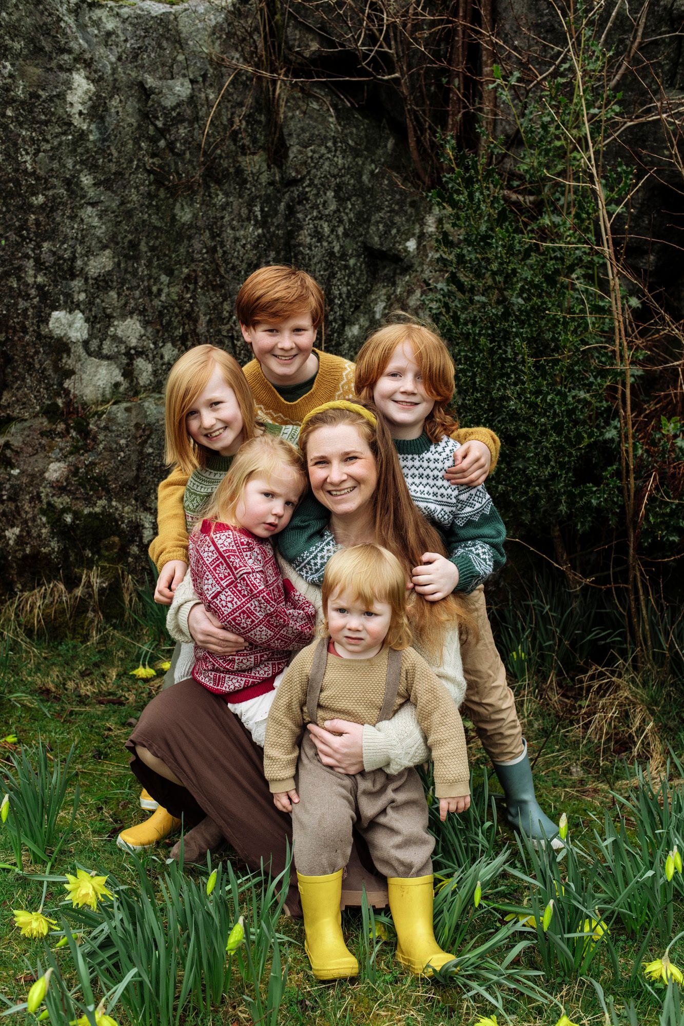 A family sits together outdoors among blooming daffodils and greenery on a spring day.