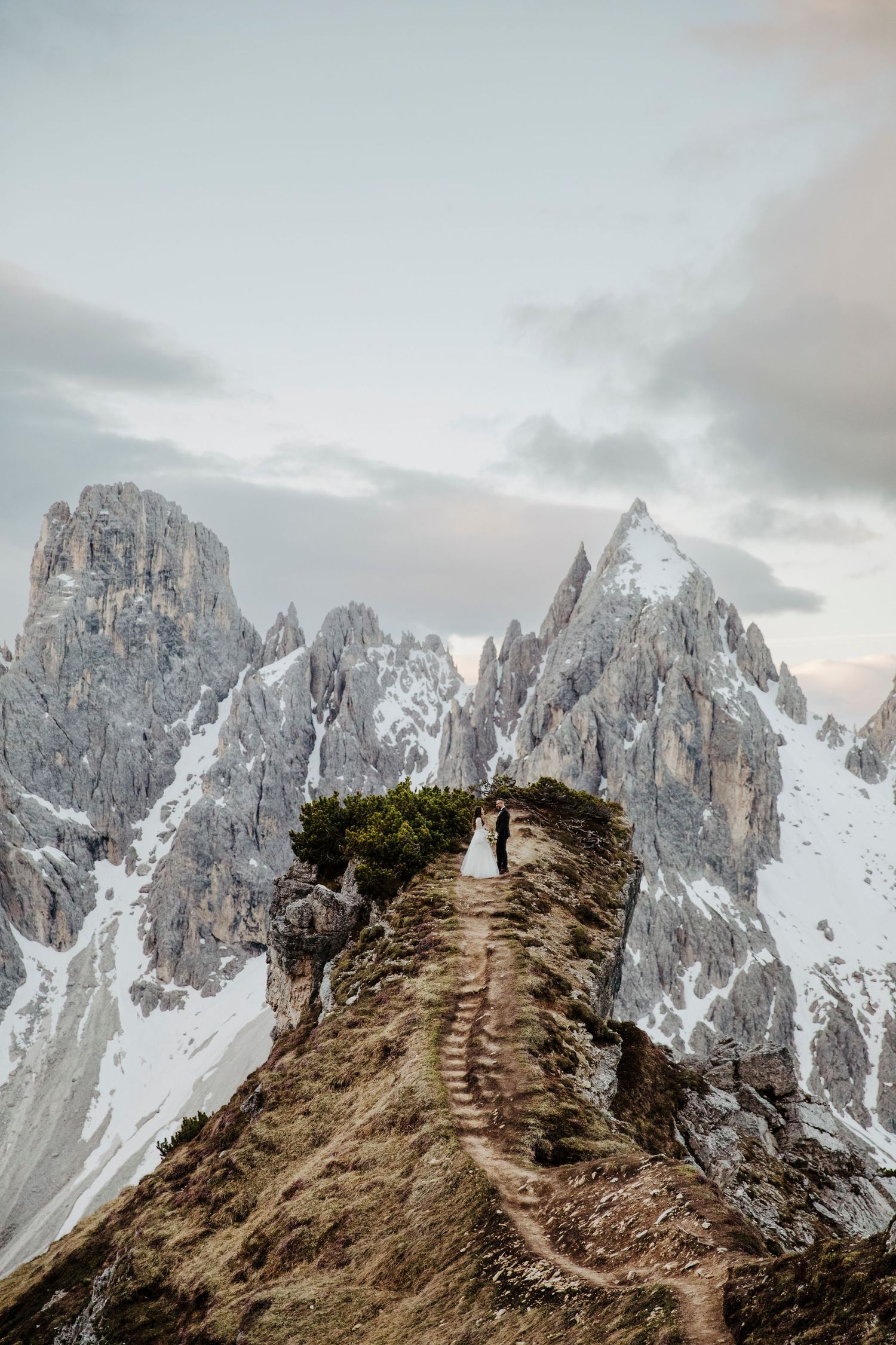 Dramatic mountain wedding photo on a narrow ridge path surrounded by snow-capped peaks.