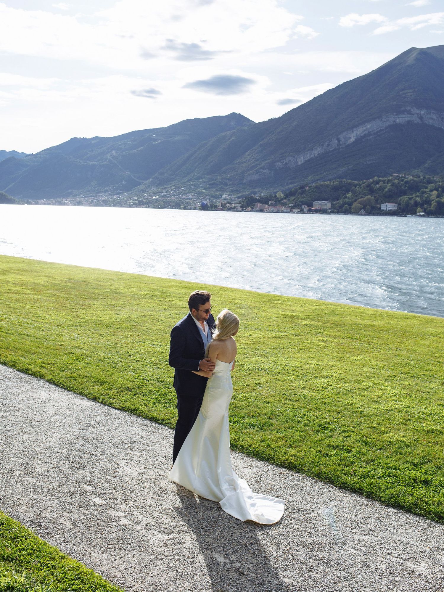 Bride and groom share a romantic moment by Lake Como, Italy, with stunning mountain views and lush green lawn.