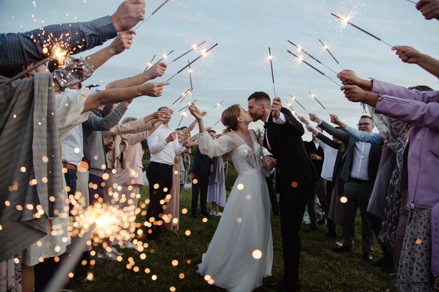 Wedding guests hold sparklers creating a magical light tunnel at dusk.