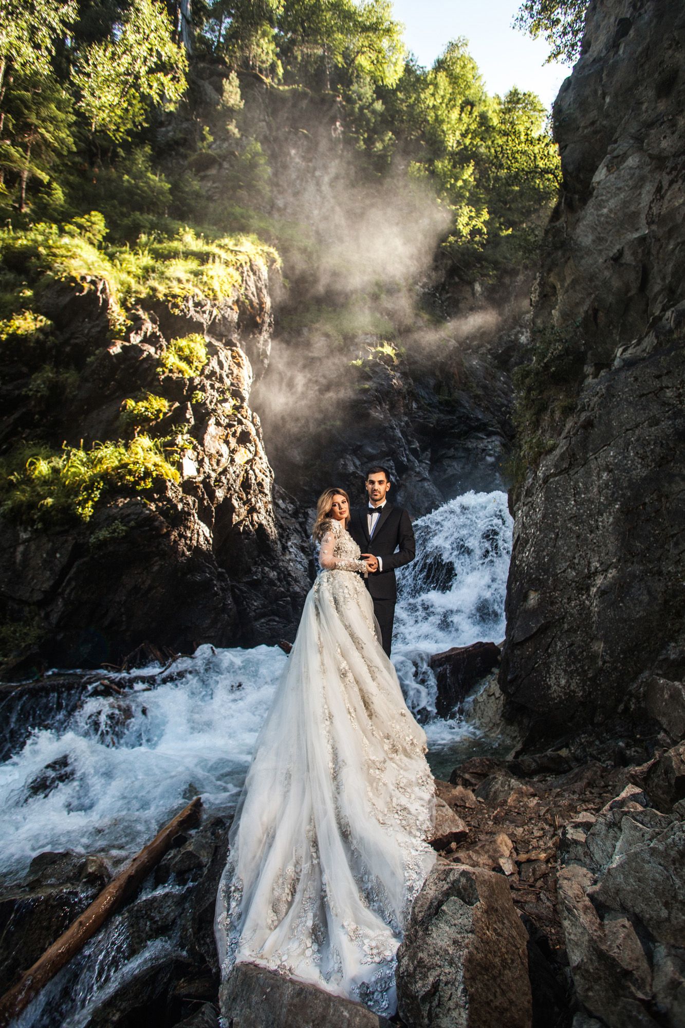 A romantic wedding photo series captured against a misty waterfall backdrop with dramatic rocky cliffs and sunlit foliage.