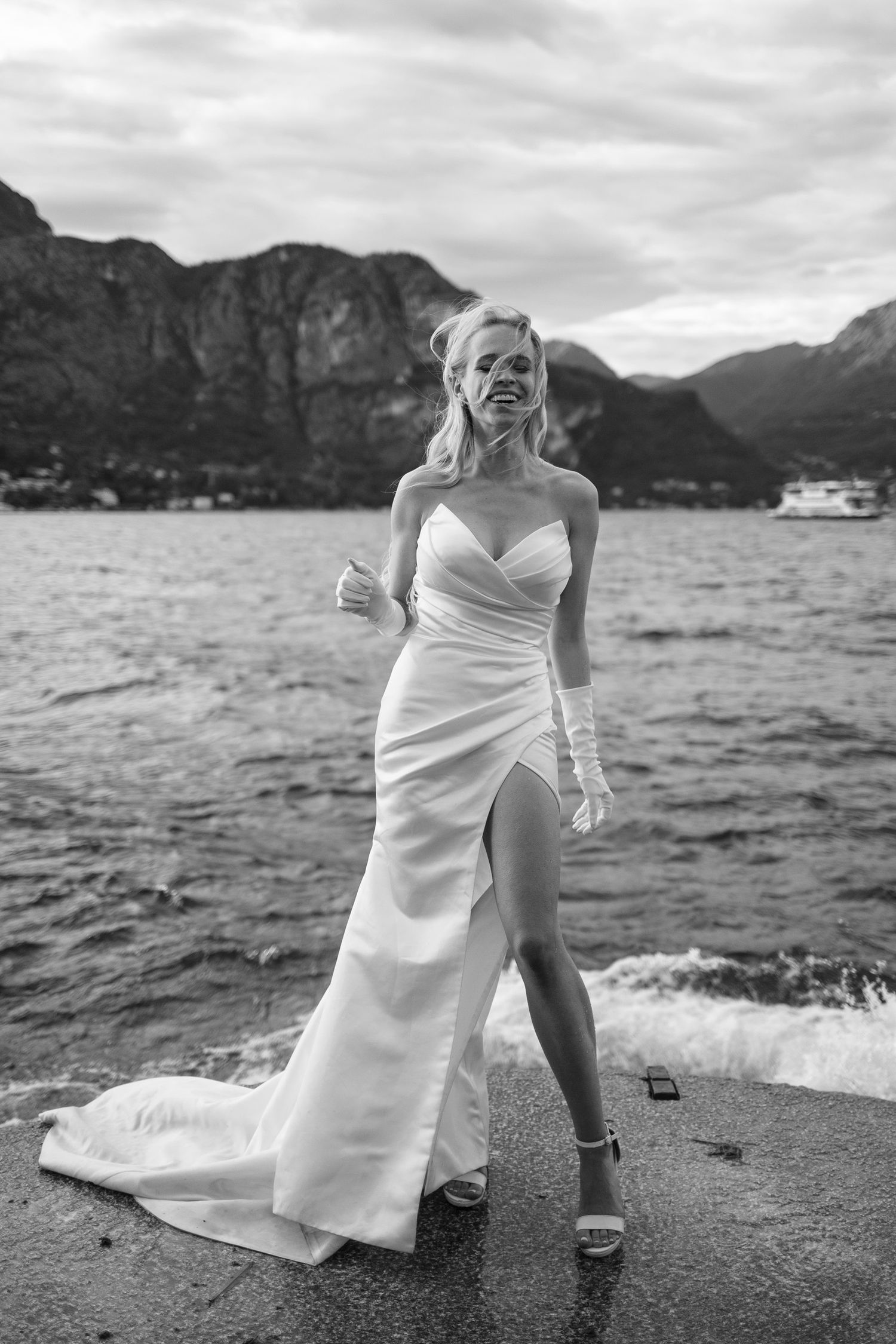 A bride in a white dress poses dramatically by a mountain lake with dramatic cloudy skies in the background.