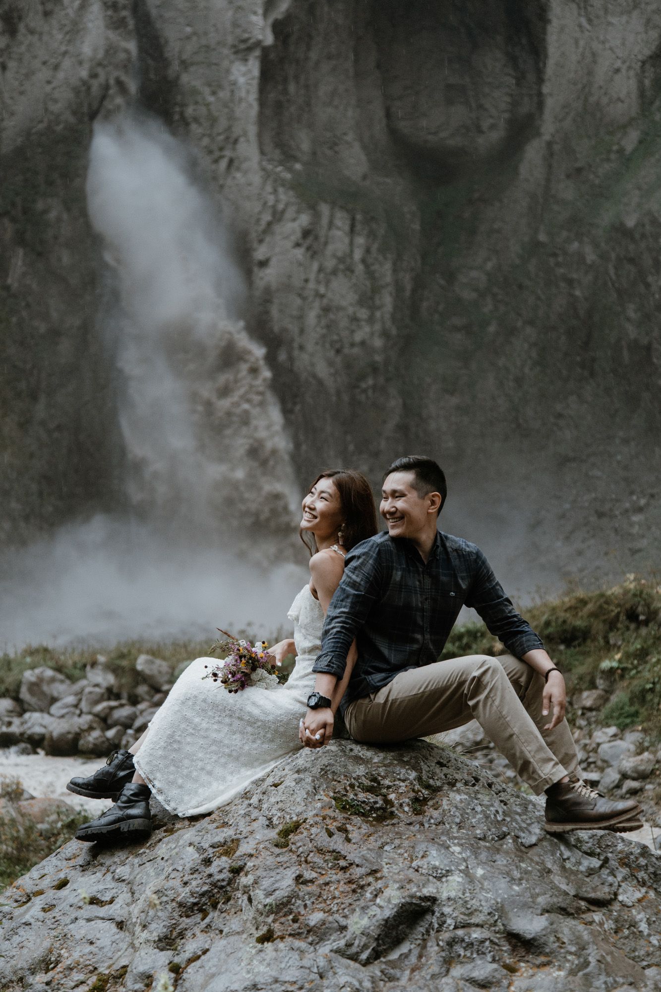 A couple sitting on rocks near a misty waterfall in a dramatic landscape.