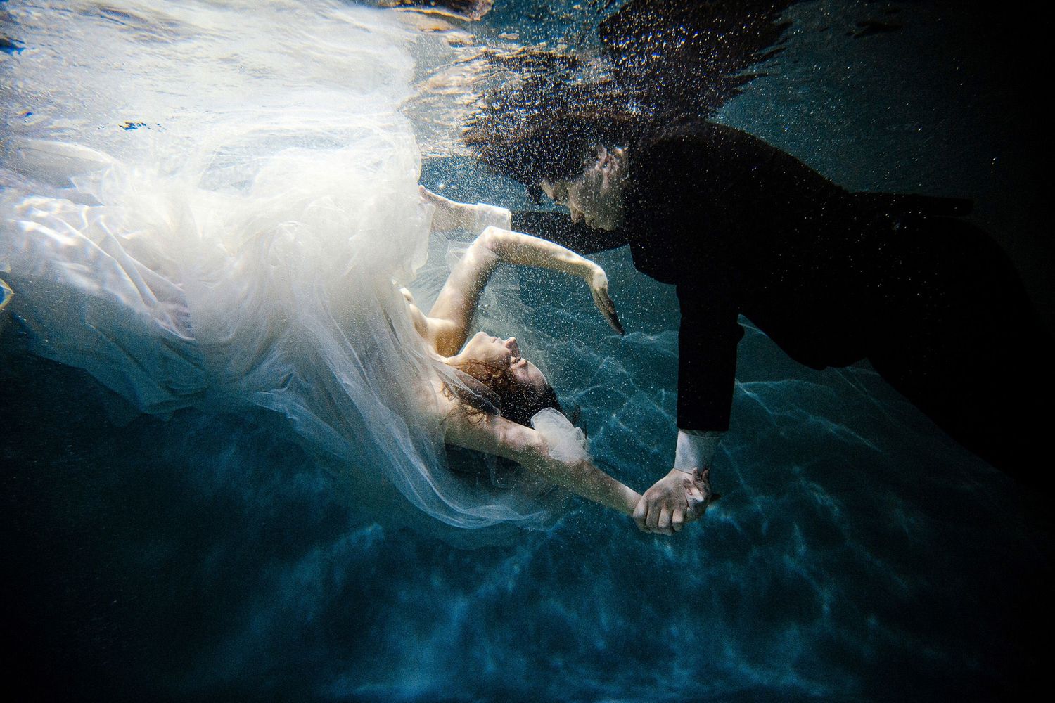 Underwater view of a swimmer in white dress floating gracefully beneath the surface in deep blue water.