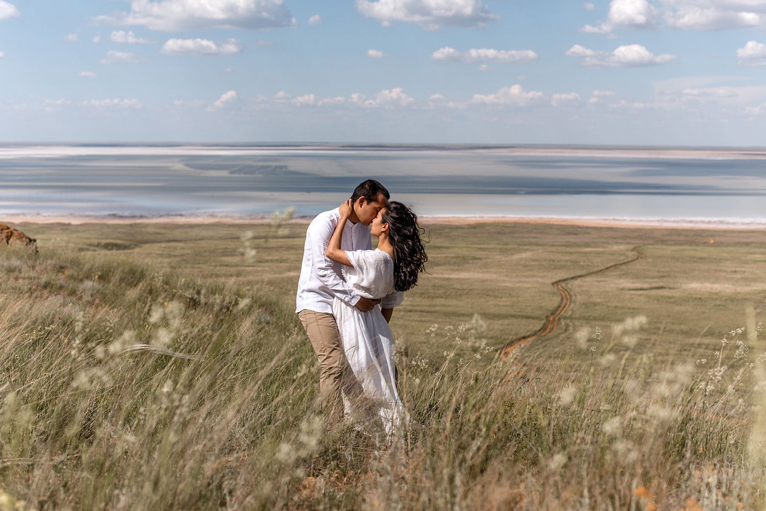 A couple in white clothing embraces on a grassy hilltop overlooking a vast ocean view on a sunny day.