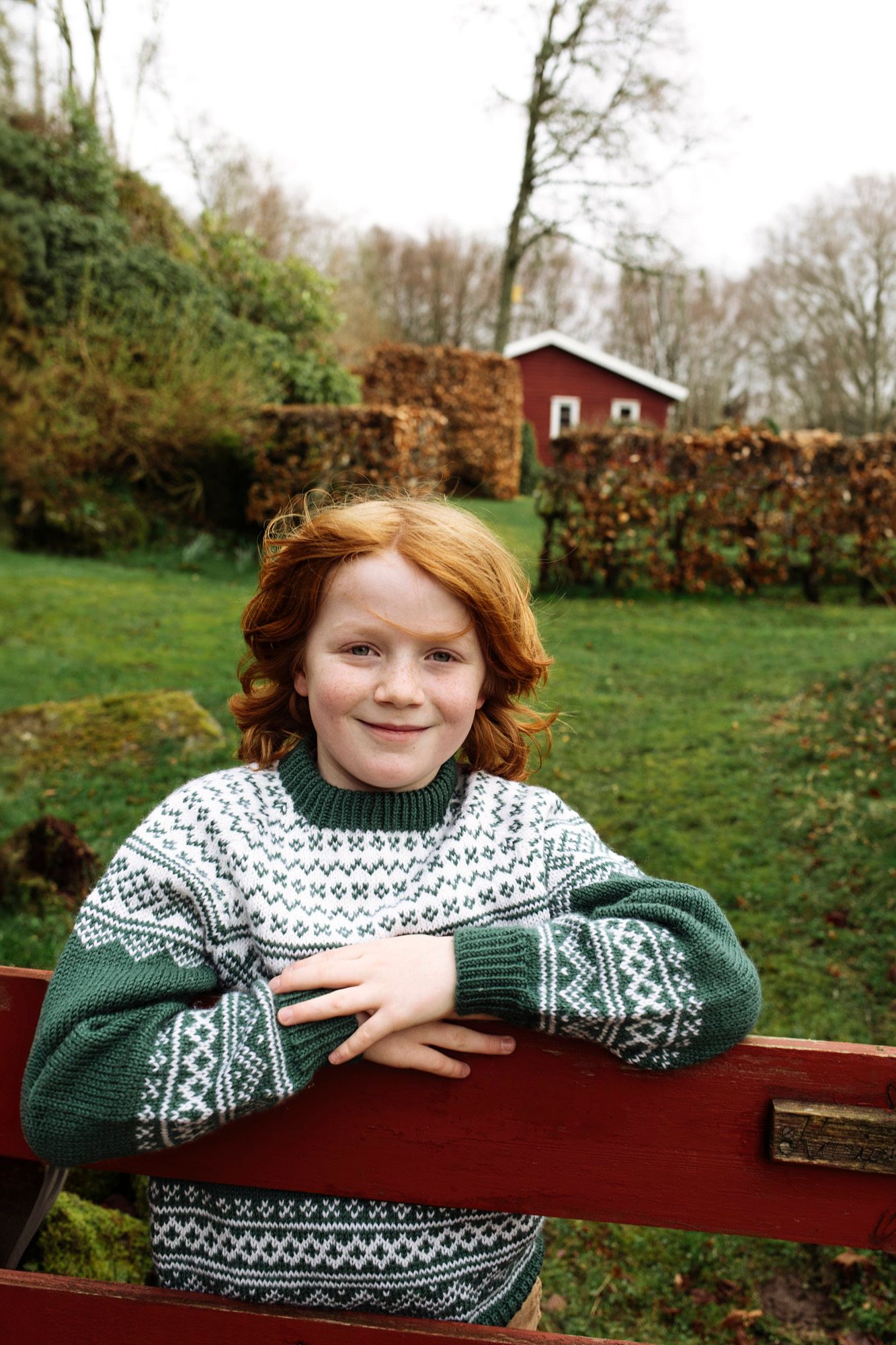 A traditional Nordic sweater is worn on a bench in front of a red cottage and autumn garden.