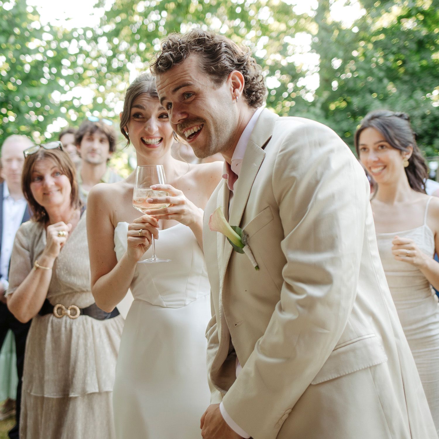 Wedding party members in cream and white attire share joyful laughter during outdoor celebration.