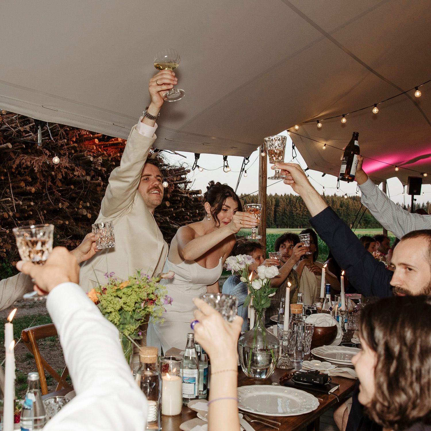 Wedding celebration under a white tent with guests raising glasses for a toast.