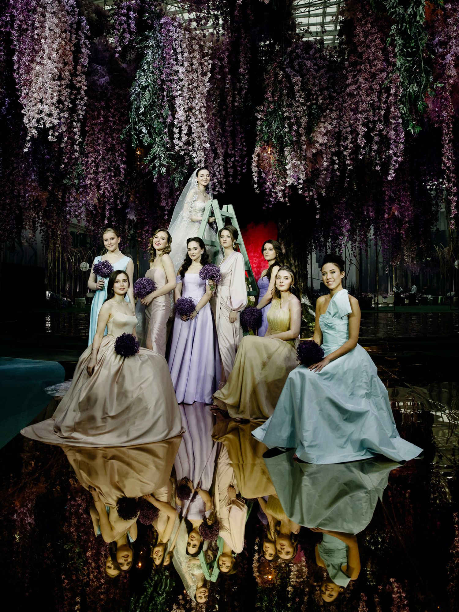 Bridesmaids in flowing pastel dresses pose beneath blooming wisteria creating a dreamy garden scene.