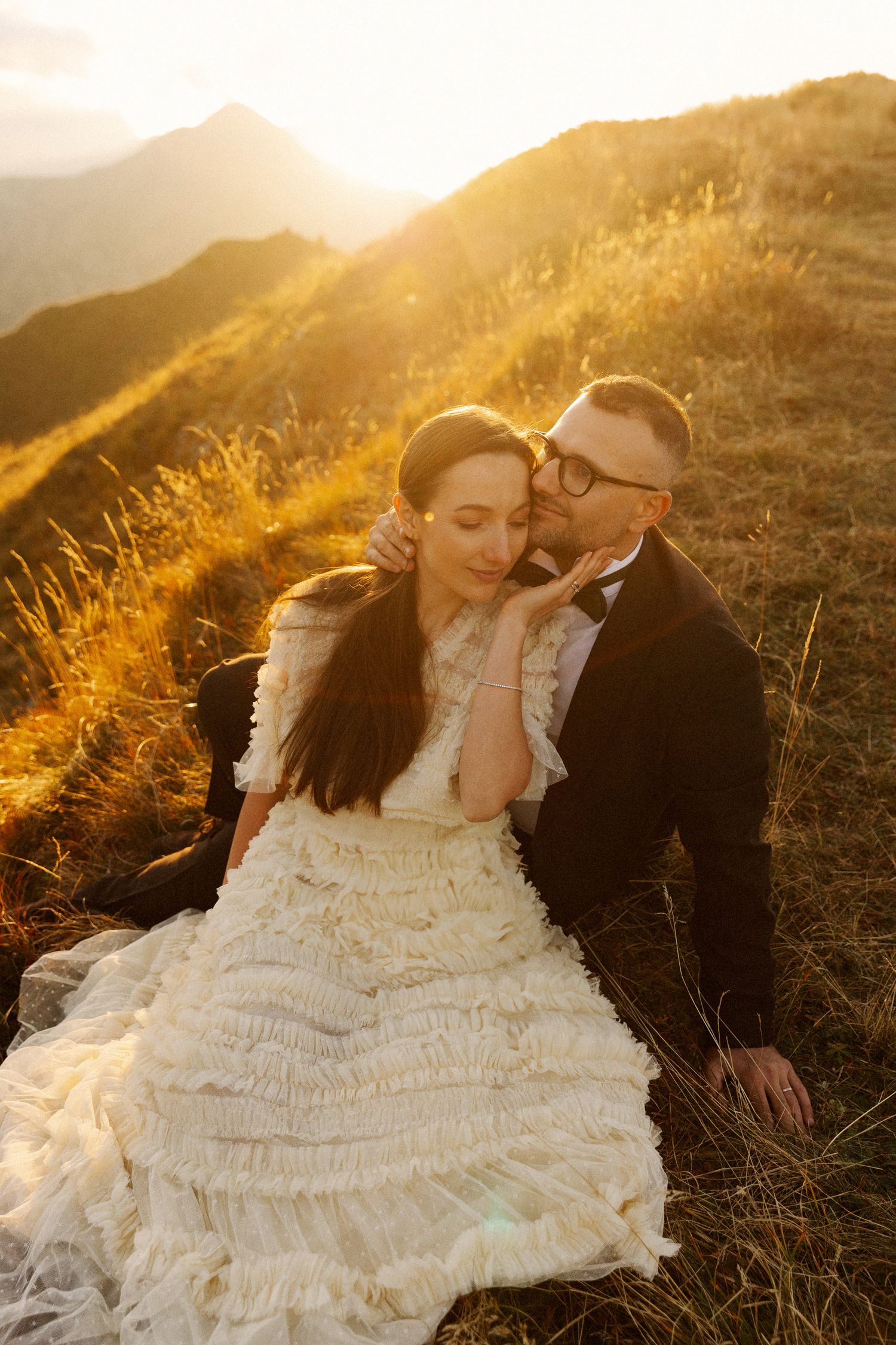 A bride and groom sit together on a sunlit mountain hillside at golden hour, sharing a romantic embrace in wedding attire.