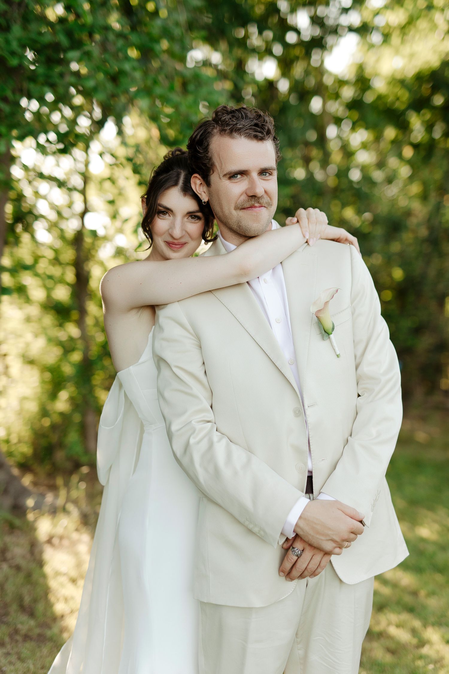 A couple in white wedding attire embraces outdoors in a sunlit garden setting with green foliage in the background.