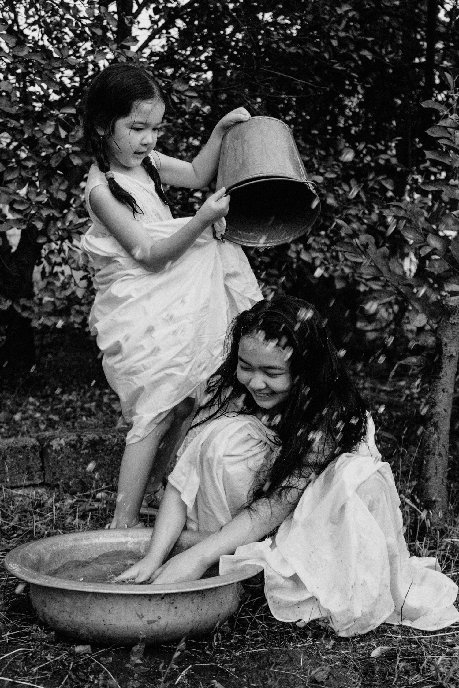 Two children in white dresses play together near a large stone birdbath in a garden setting, captured in black and white.