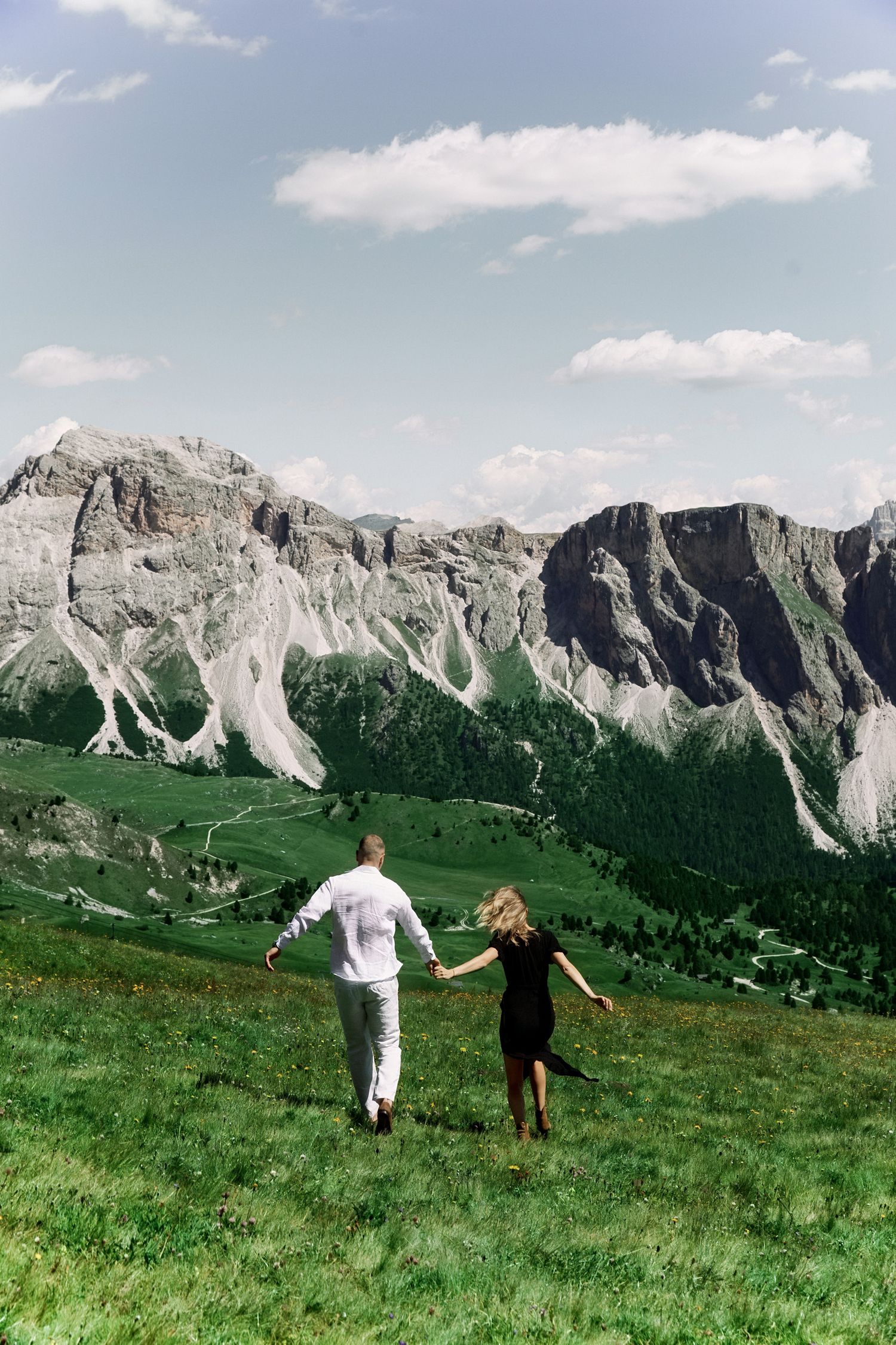 Couple holding hands walking on green alpine meadow with dramatic Dolomite mountain peaks and blue sky backdrop.