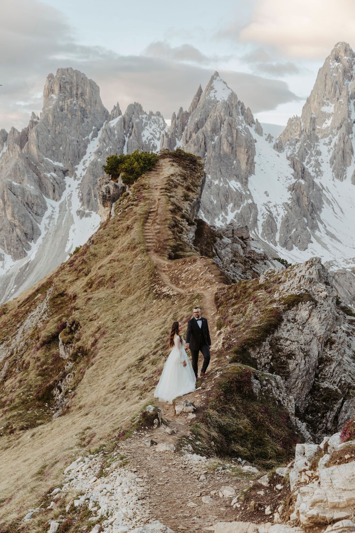 Hikers traverse a narrow mountain ridge trail with dramatic jagged Dolomite peaks rising in the misty background.