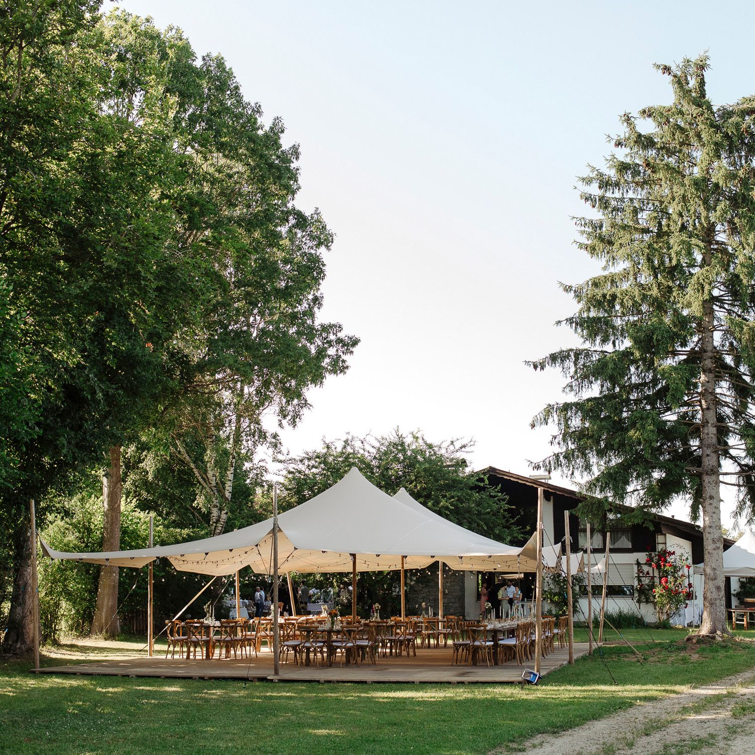 Outdoor wedding reception setup with white stretch tents and rustic wooden furniture arranged on a grassy lawn.