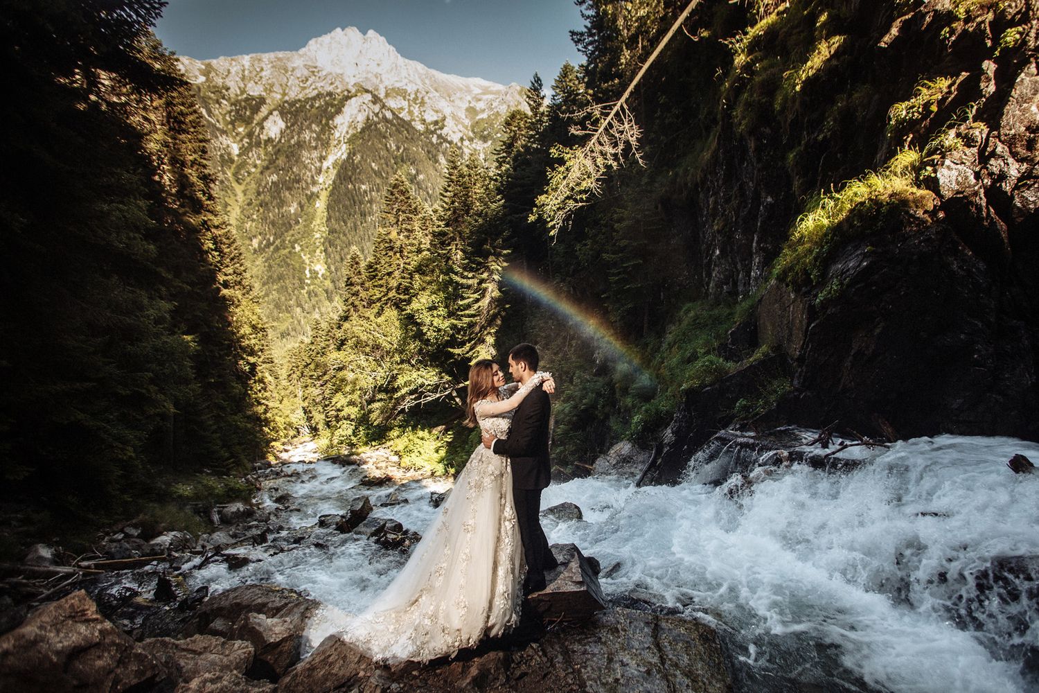 A couple poses in front of a rushing waterfall surrounded by lush forest in a mountain landscape.