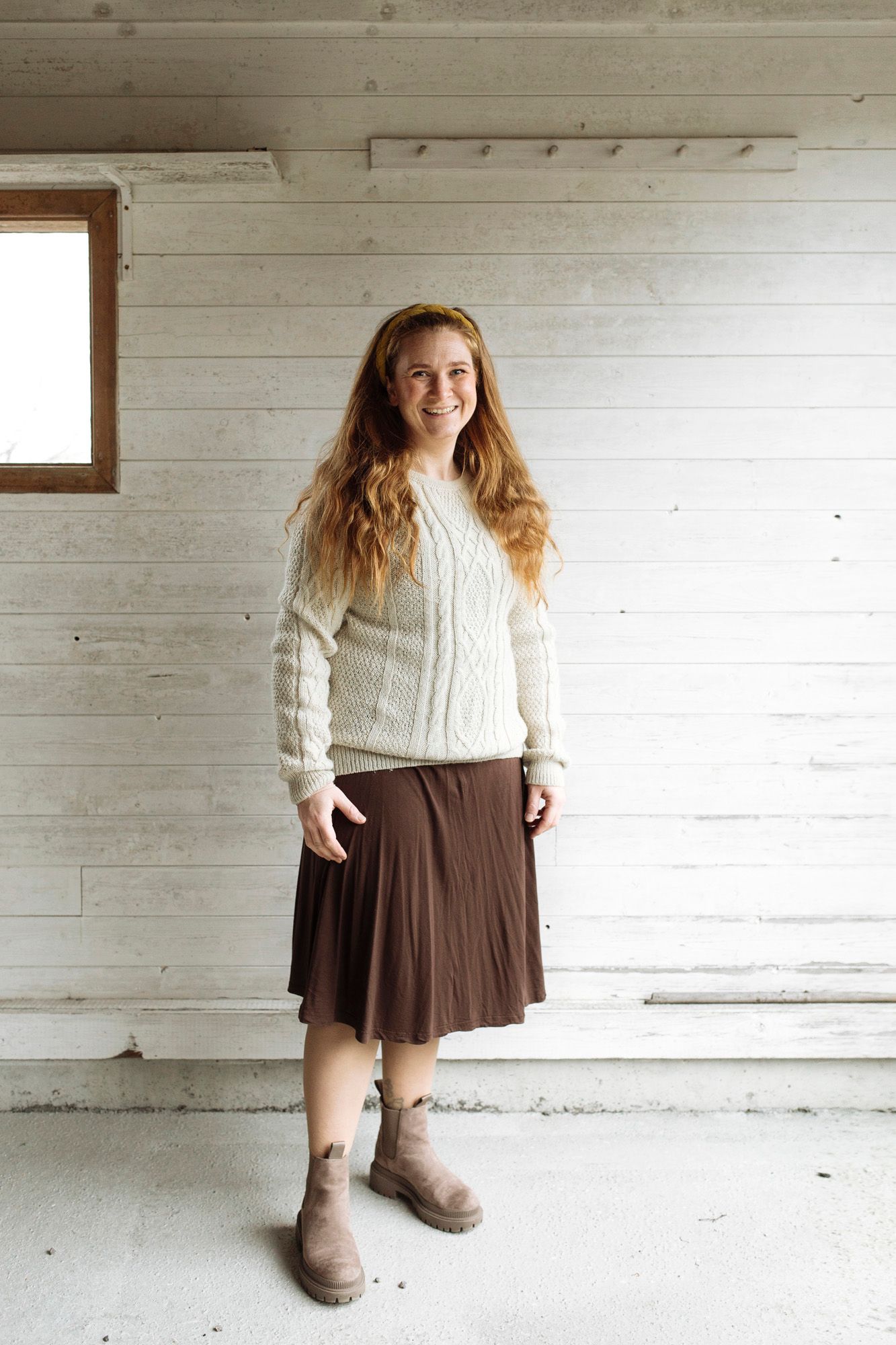 Classic white cable knit sweater paired with brown pleated skirt and boots in rustic indoor setting.