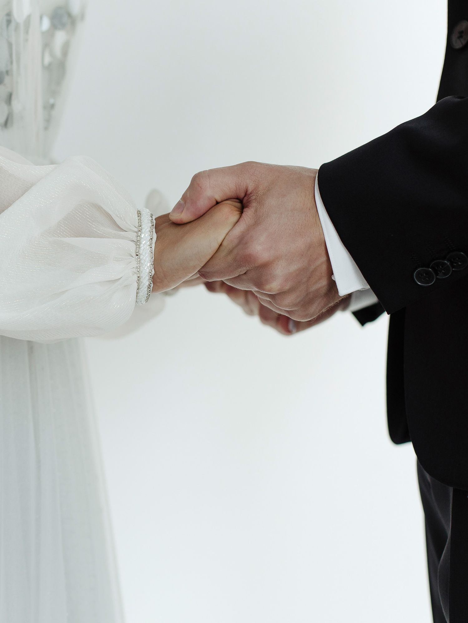 Close-up of bride and groom holding hands during wedding ceremony, with white dress and black suit visible.