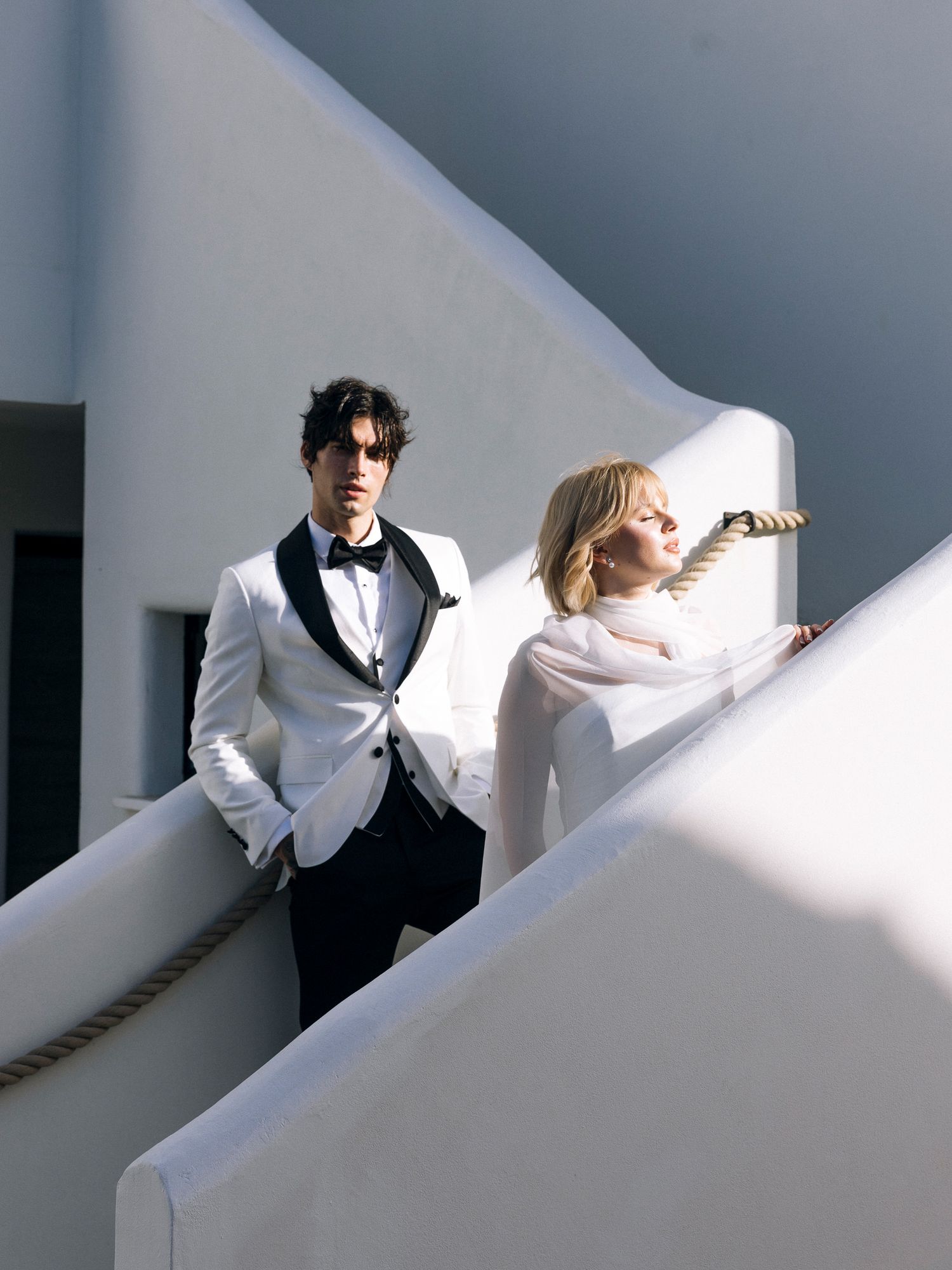A person in a black suit and white shirt stands against a white wall creating dramatic angular shadows.
