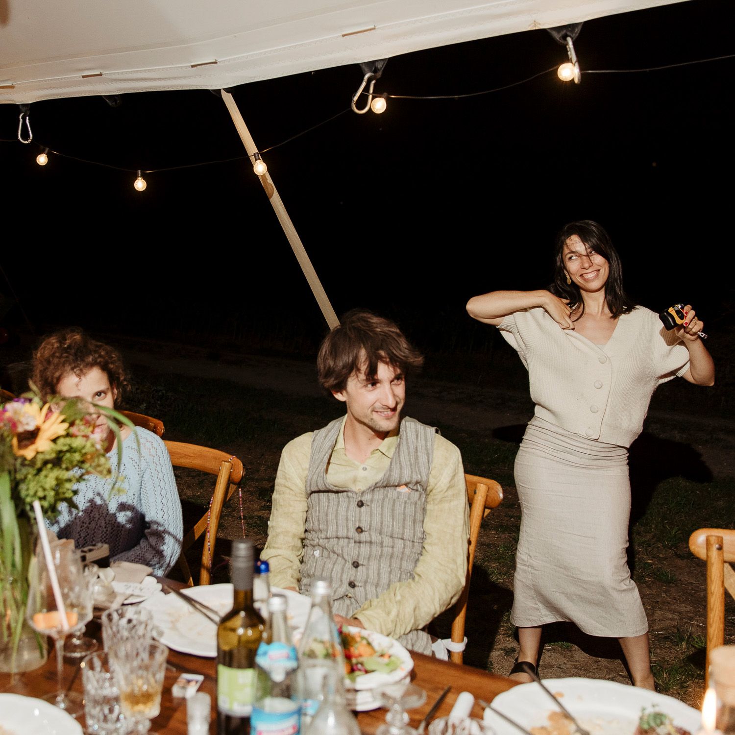 People enjoying an outdoor dinner party at night under string lights with food and drinks on a long table.