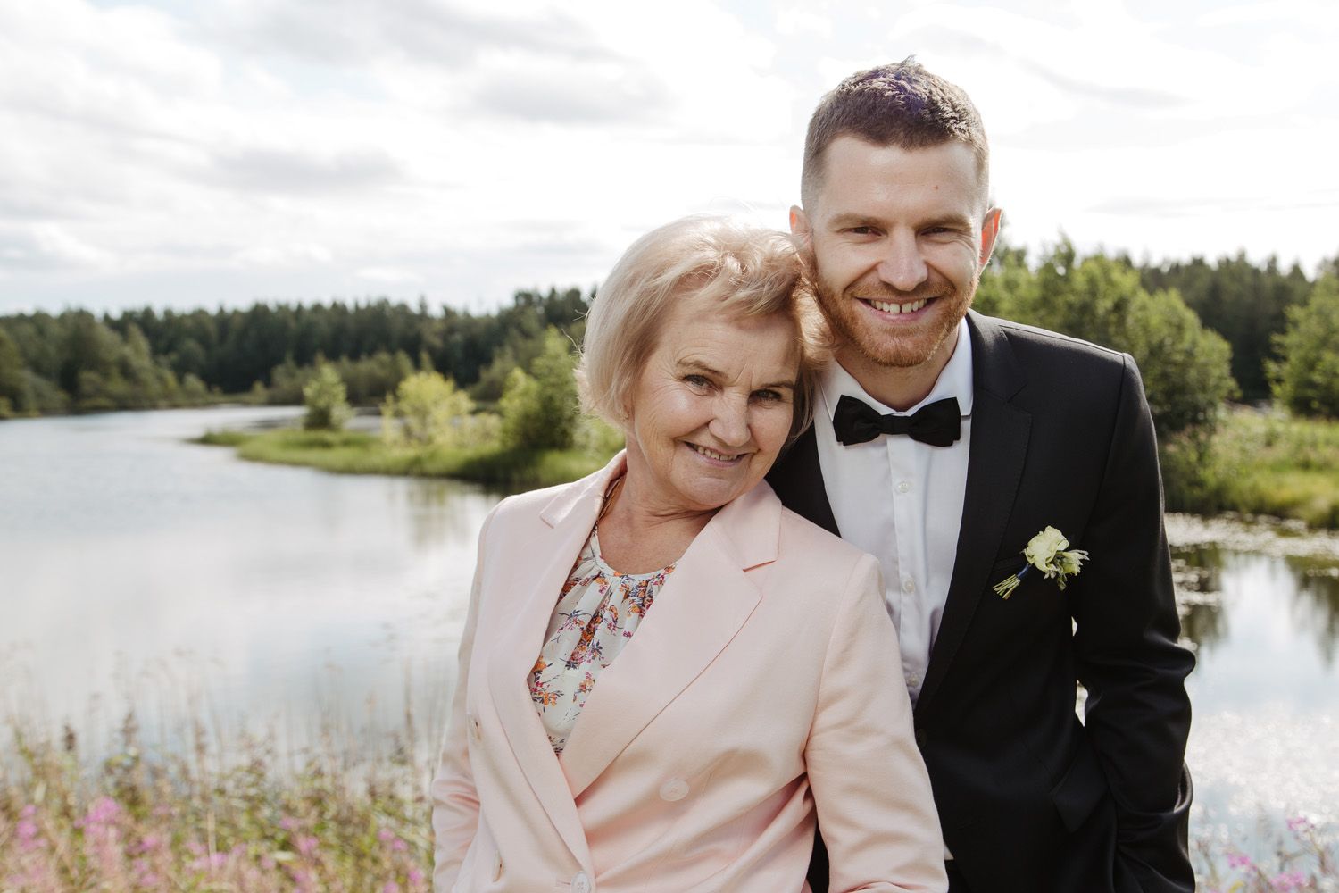 Couple in formal attire posing by lakeside with trees in background.