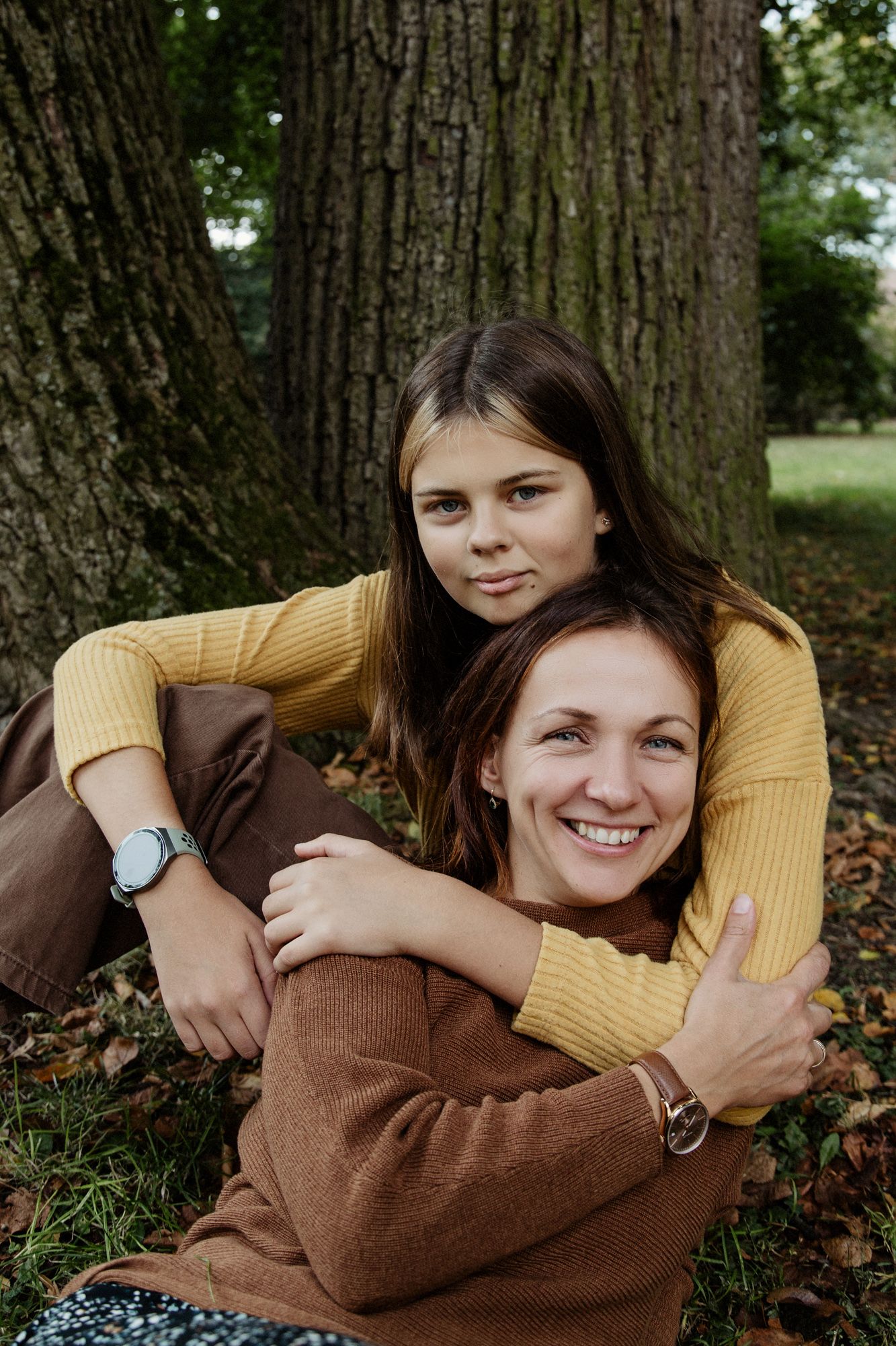Two people in brown and yellow sweaters share a warm embrace while sitting among fallen autumn leaves near a tree.