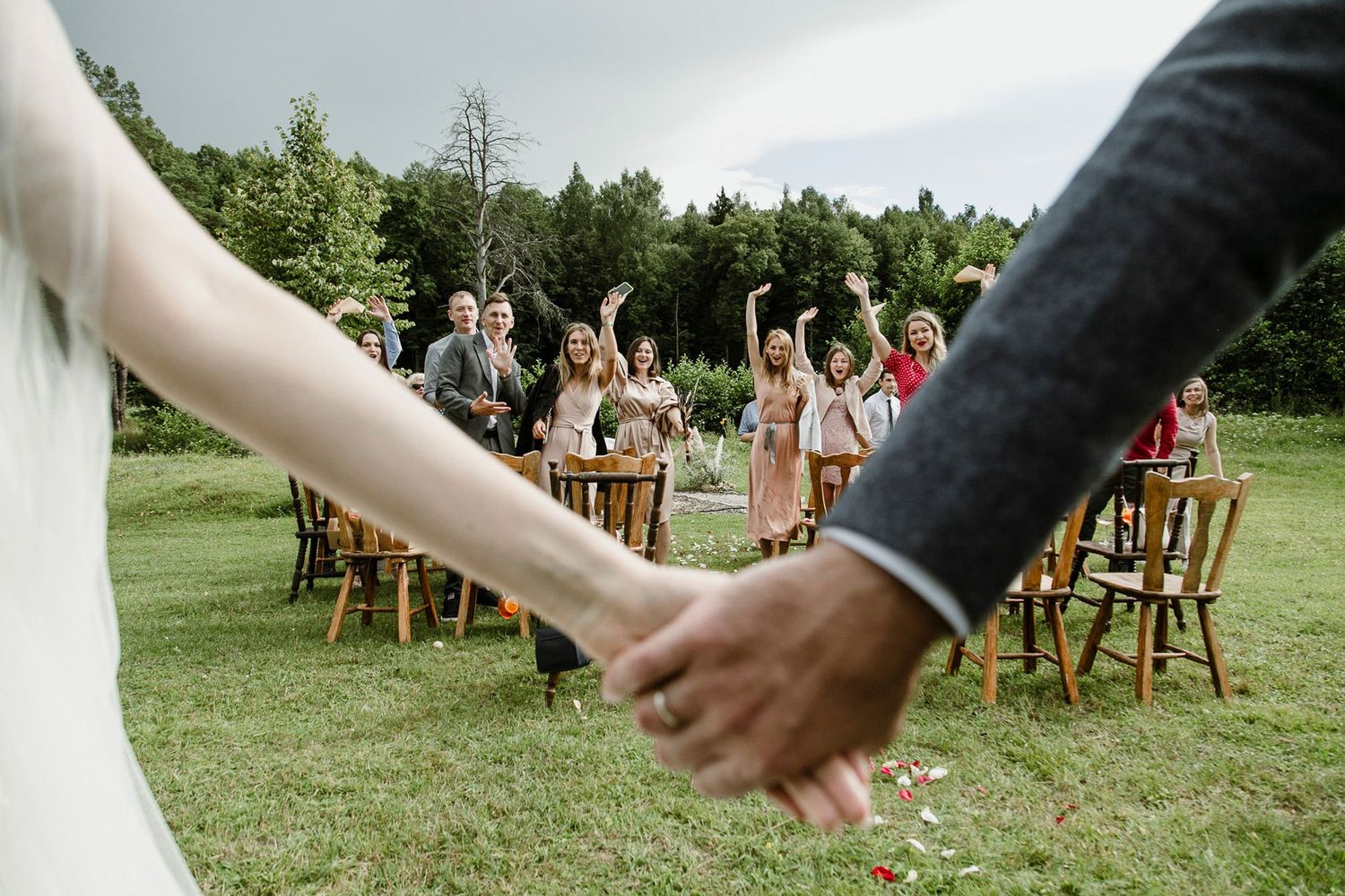 A couple holds hands during an outdoor wedding ceremony with wooden chairs and guests on a green lawn.