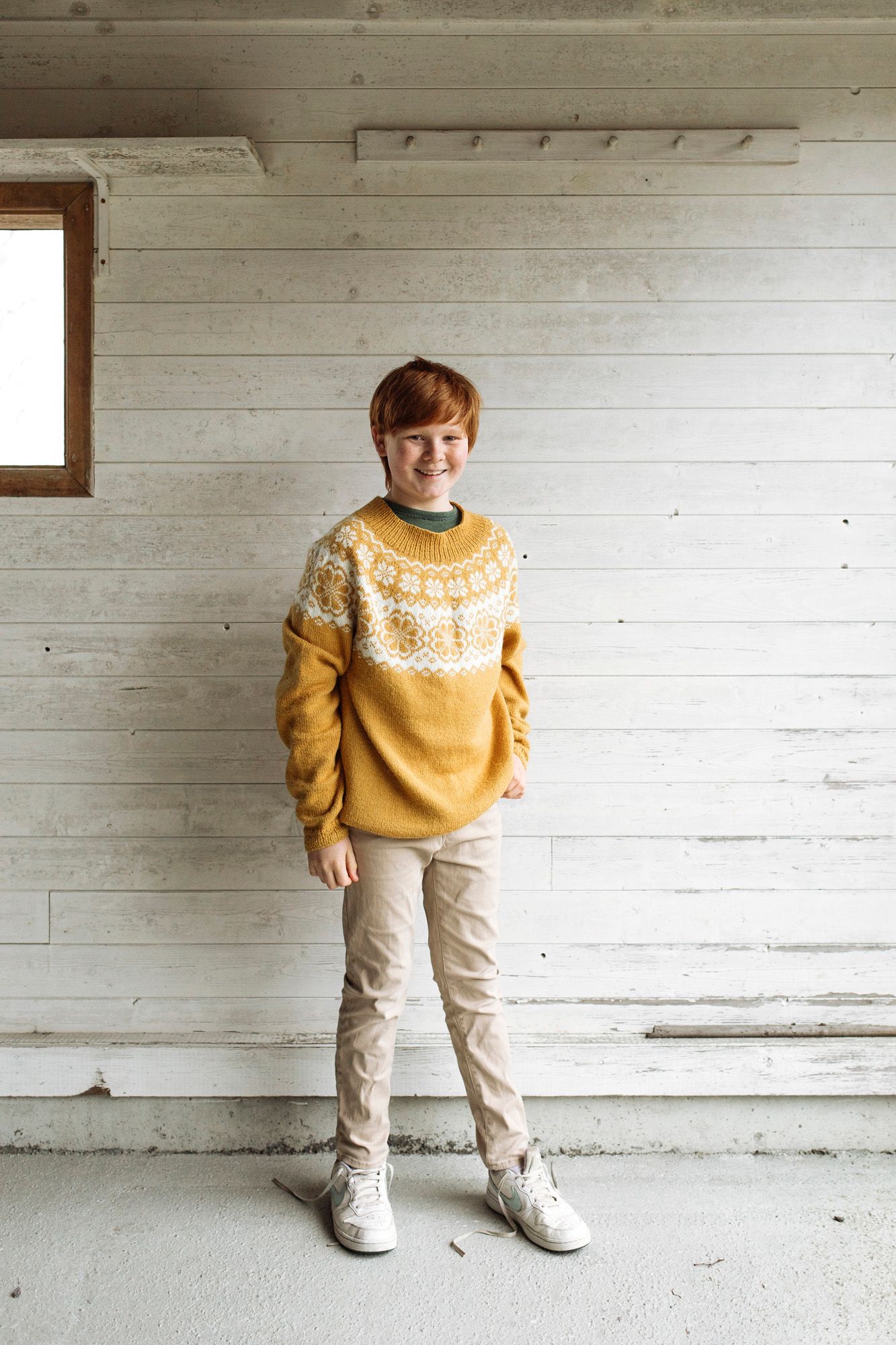 Modern casual look with mustard fair isle sweater and beige pants against white wooden wall.
