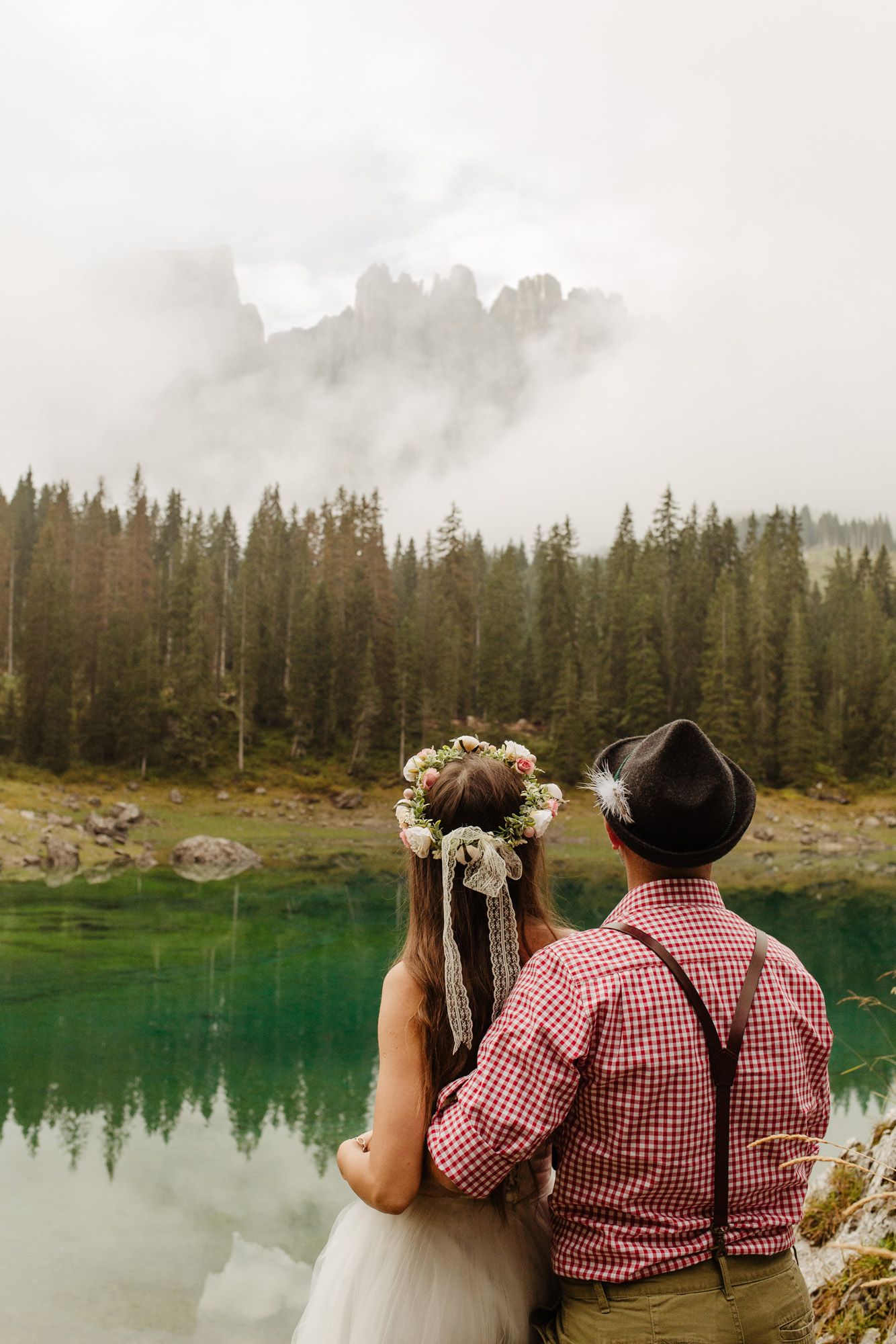 Couple in traditional red and white checkered outfits admiring a serene turquoise alpine lake surrounded by evergreen forest.