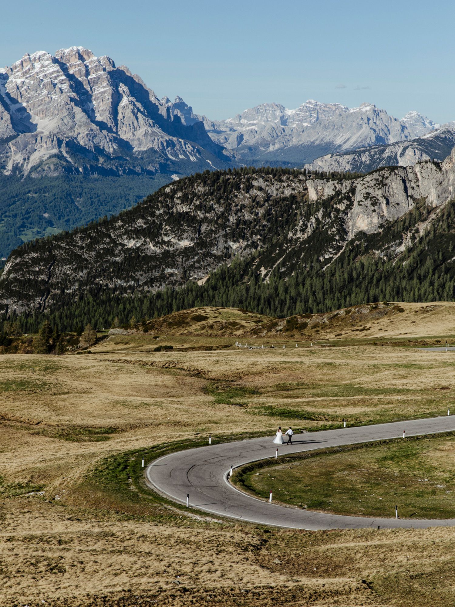 Winding mountain road curves through scenic alpine landscape with rugged peaks and green forests in the background.