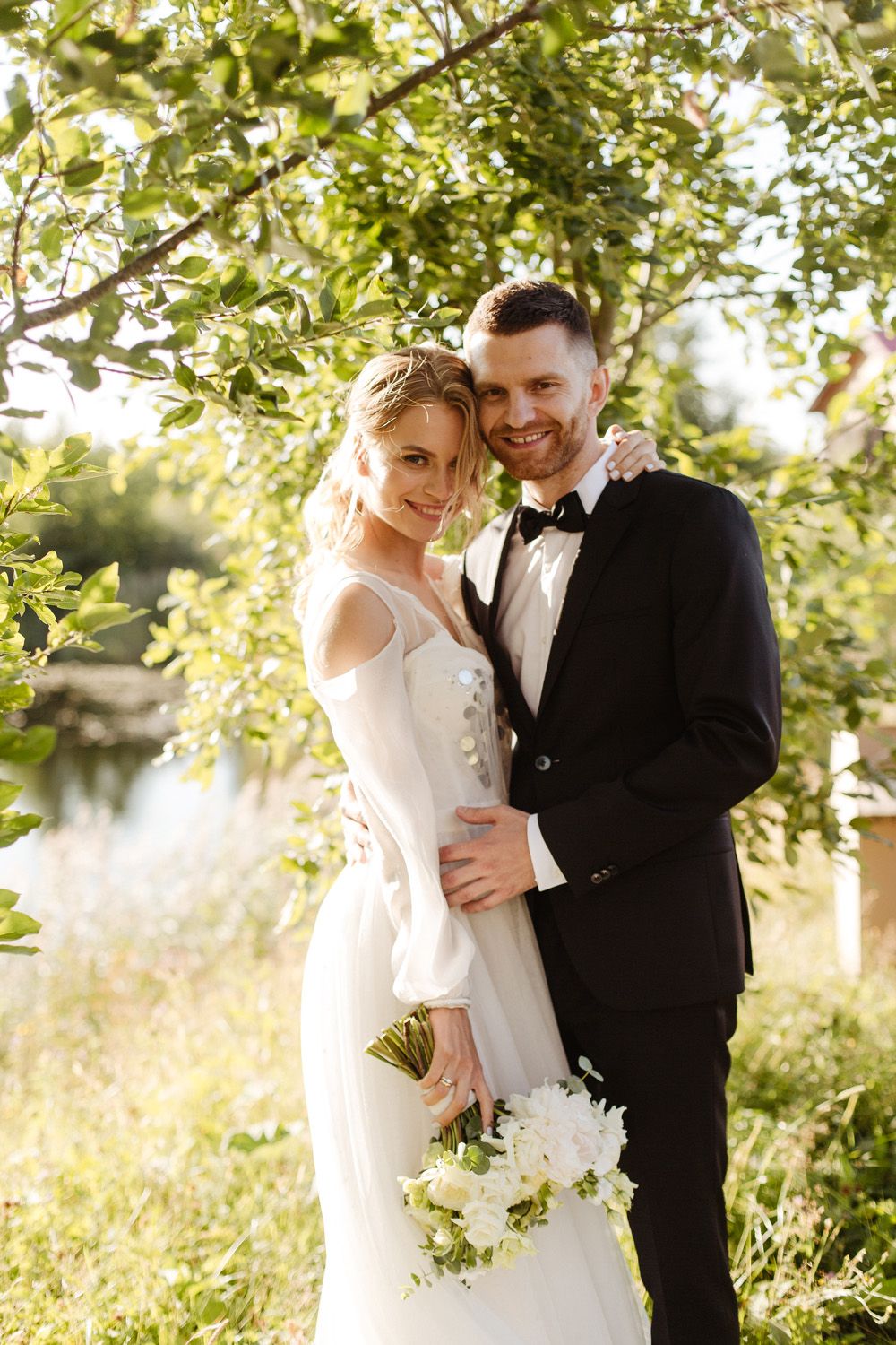 Newlywed couple embraces under sunlit trees in an outdoor garden setting with white floral bouquet.