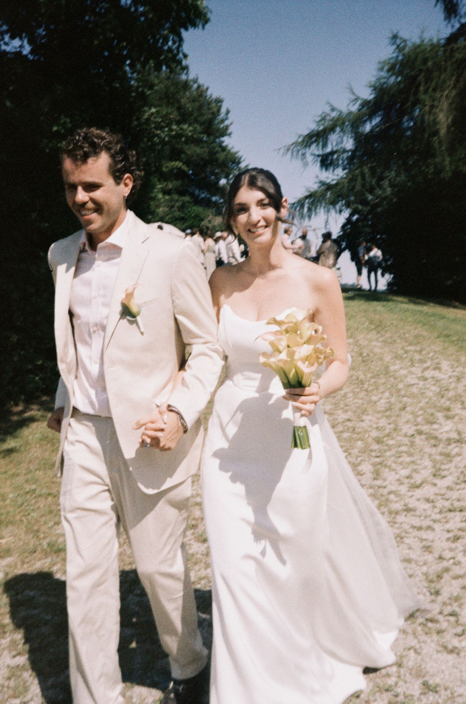 A happy couple walks together on a sandy beach path wearing elegant white wedding attire.