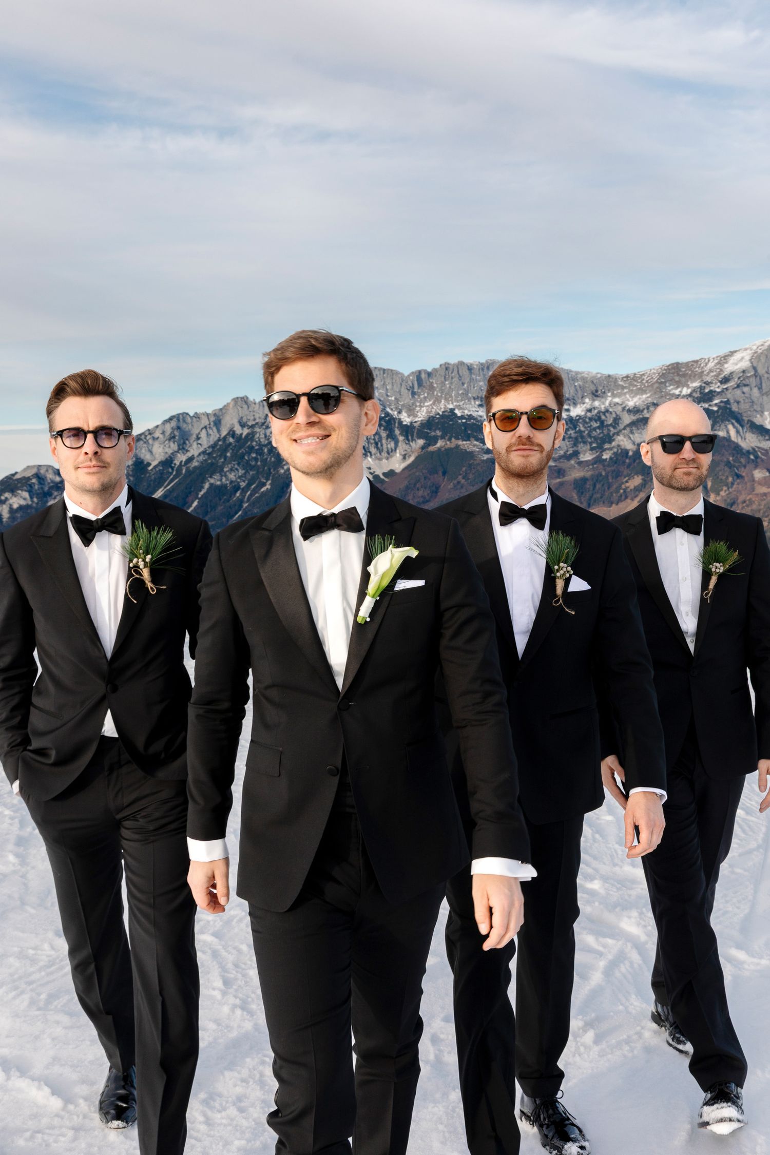 A group of groomsmen in black tuxedos and bow ties pose together against a snowy mountain backdrop.