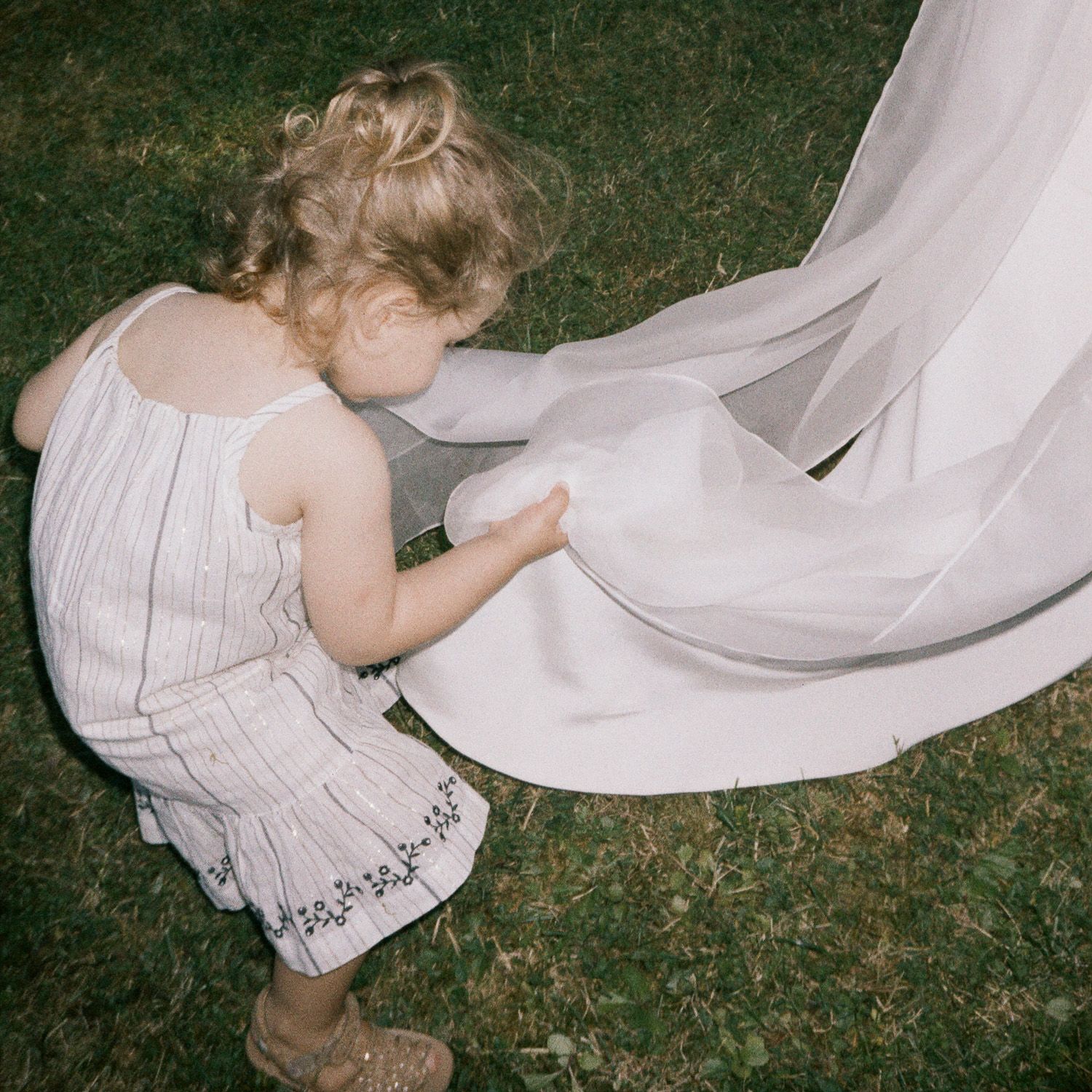 A small child in white dress plays with flowing fabric on grass during an outdoor event.