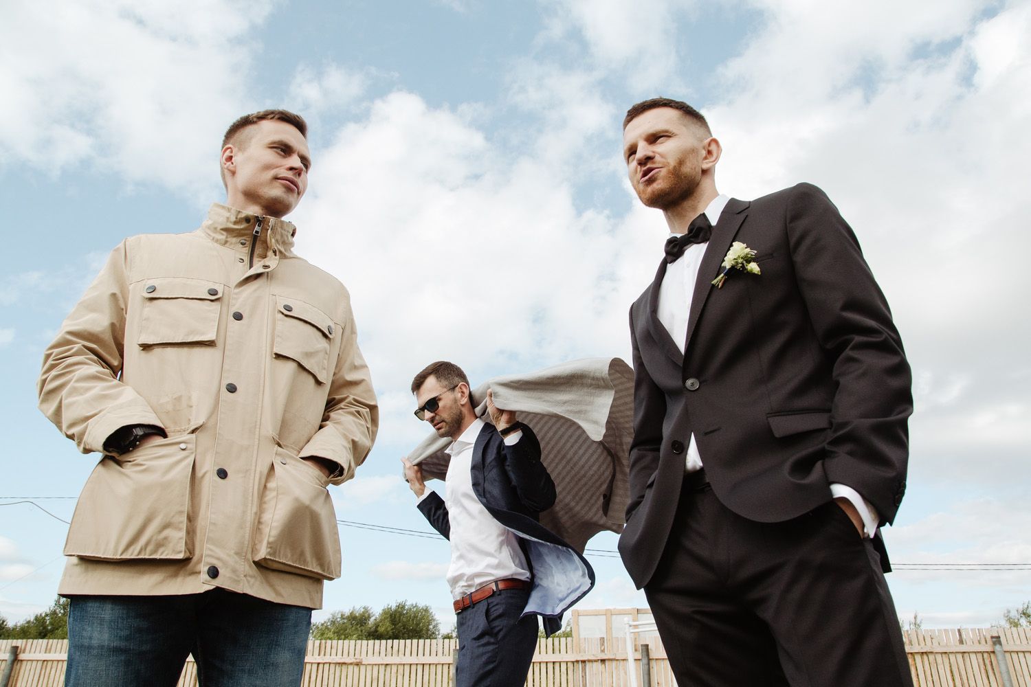 Three men in formal attire standing outdoors against cloudy sky.