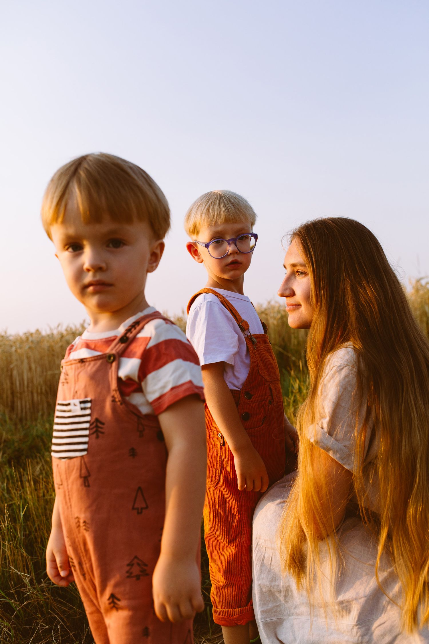 Children and adult with long hair stand together in a grassy field at sunset wearing casual summer clothing.
