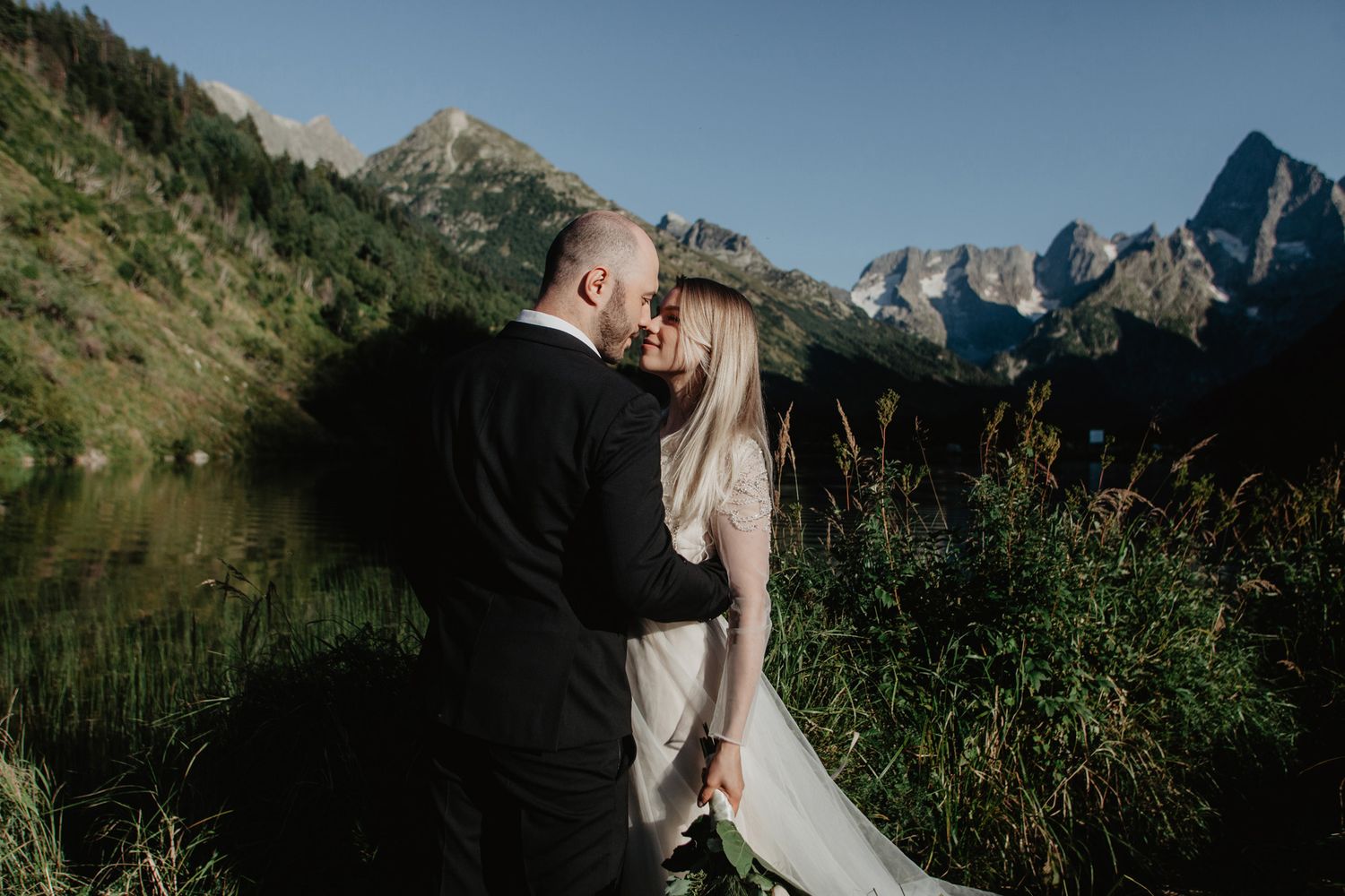A romantic wedding couple embraces against a dramatic mountain landscape with evergreen trees.