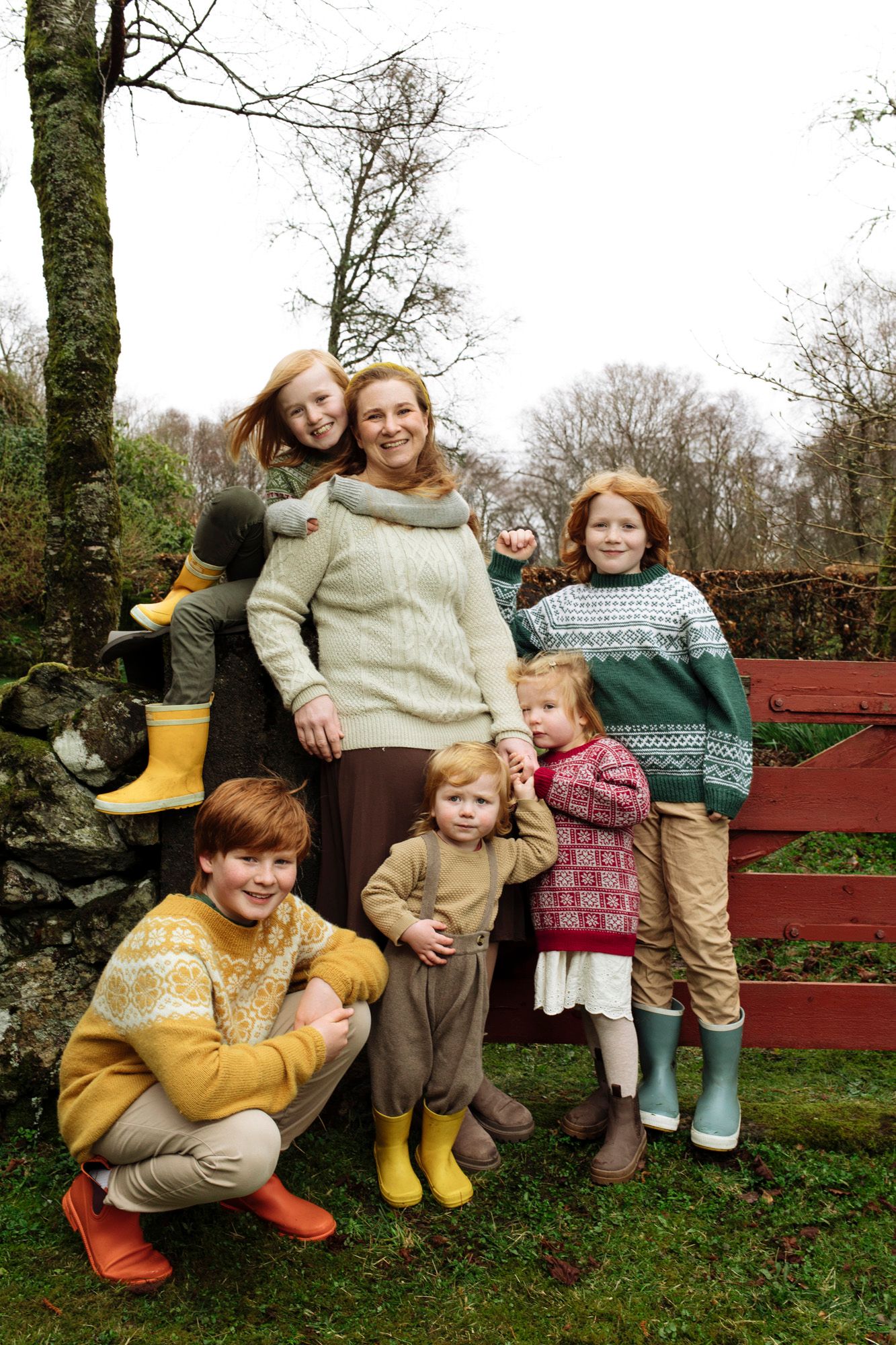 Group wearing coordinated knitwear and colorful boots posed by red wooden fence.
